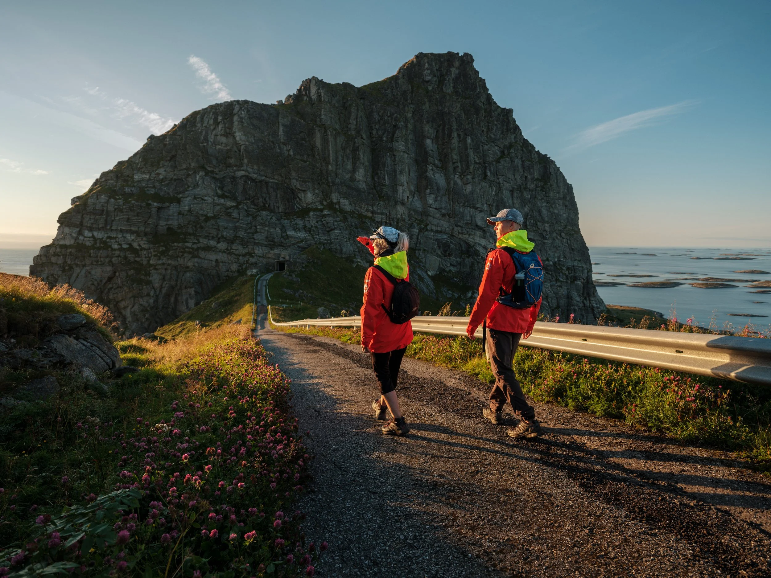 Two hikers in red jackets and backpacks walking on a dirt trail near a large rocky mountain at sunset, with sea and sky in the background.
