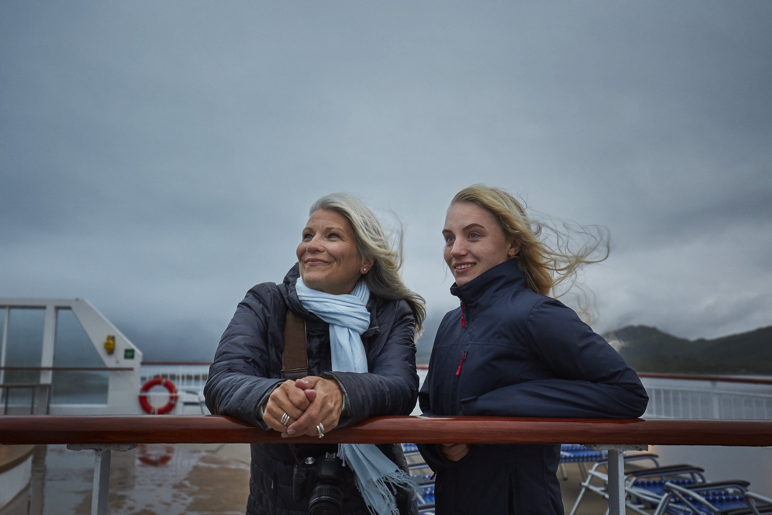 Two women standing on a ship's deck, smiling and enjoying the wind, with cloudy sky and mountains in the background.