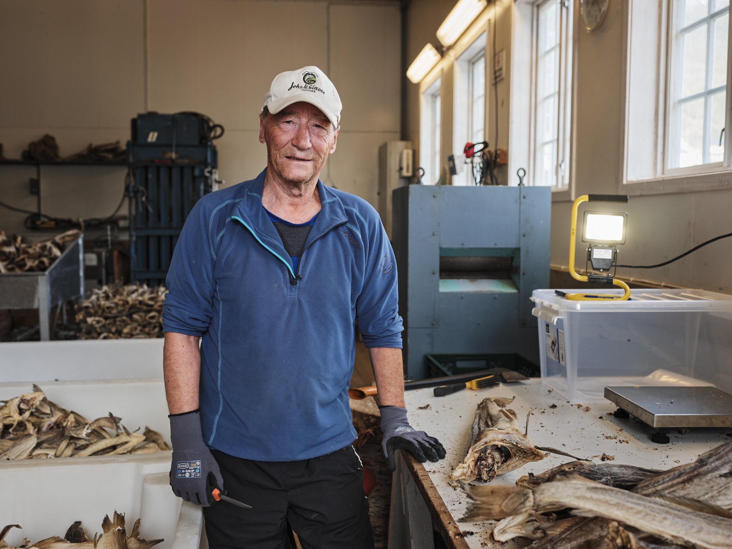 An older man in a blue jacket and white cap stands in a seafood processing facility, holding a knife, with fish and fish heads on a table in front of him.