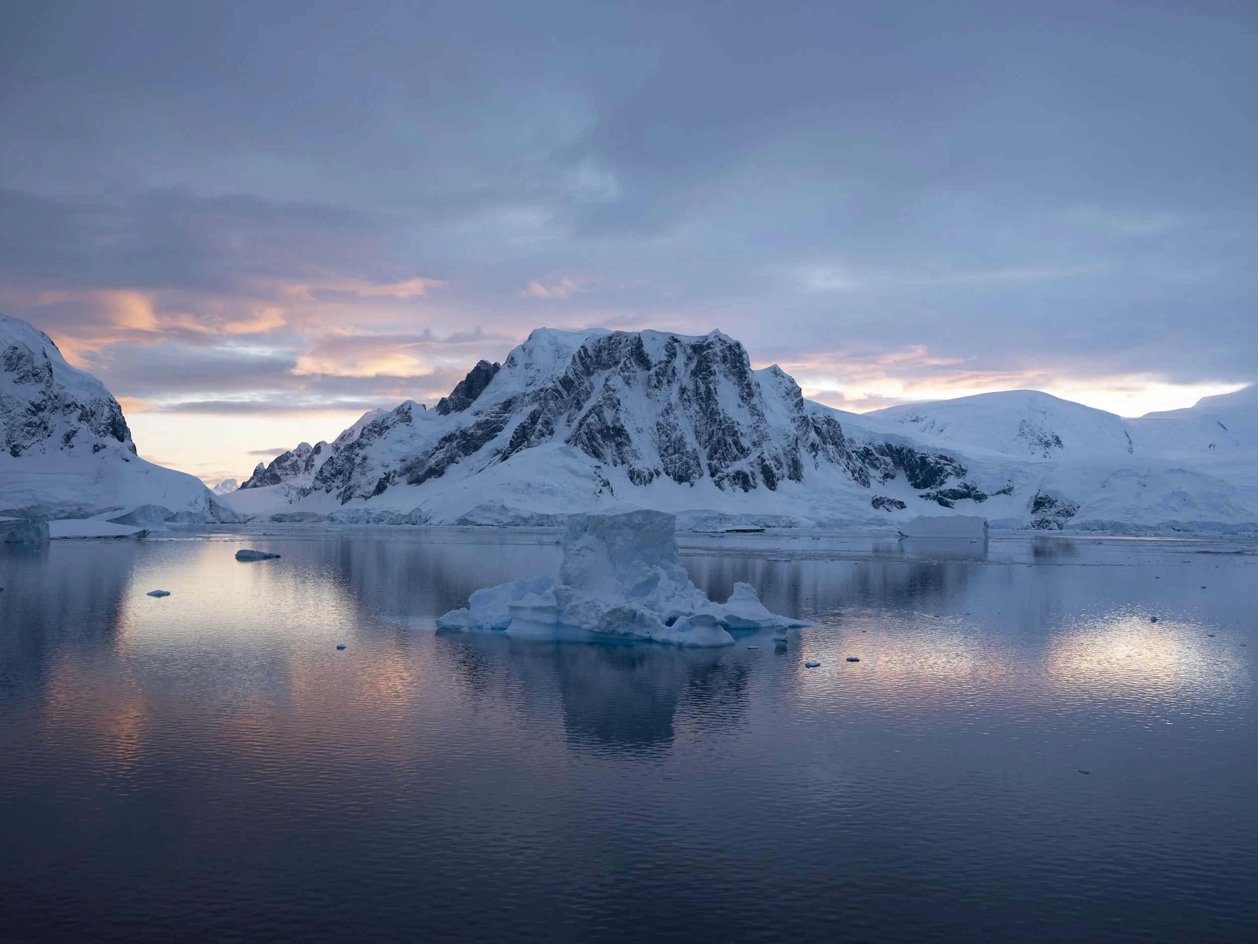 Snow-covered mountains reflecting in calm icy water with icebergs and a colorful sky.