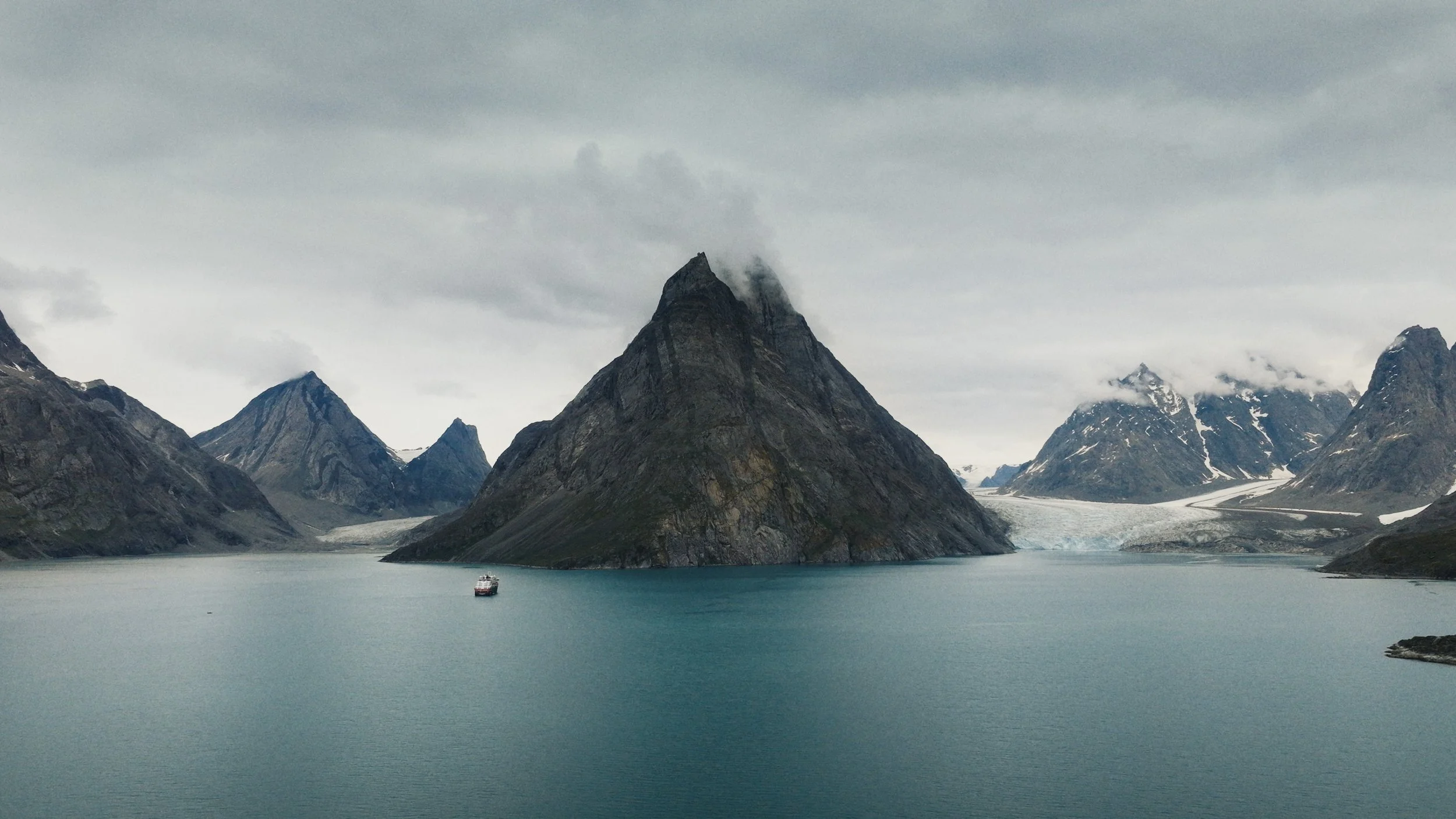 A fjord with towering mountain peaks, some snow-capped, under an overcast sky. A boat floats on the calm, teal water near the base of a large, pyramid-shaped mountain. Greenland. Expedition.