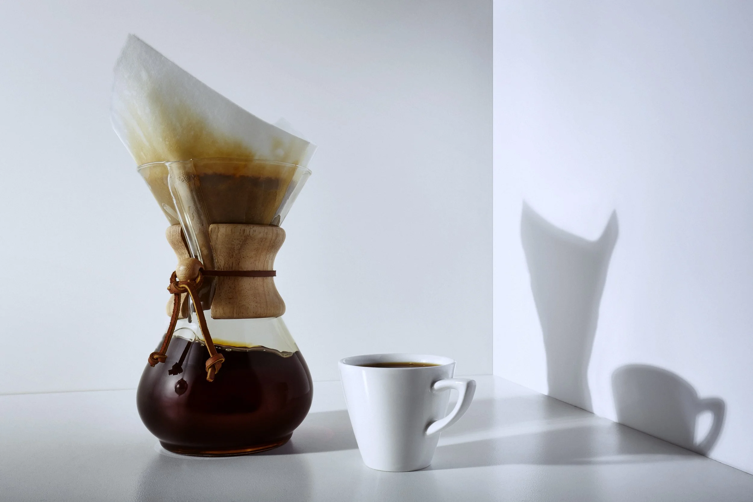 A Chemex coffee maker with a paper filter and brewed coffee, placed on a white surface next to a white coffee cup with coffee inside, casting shadows on a white background.