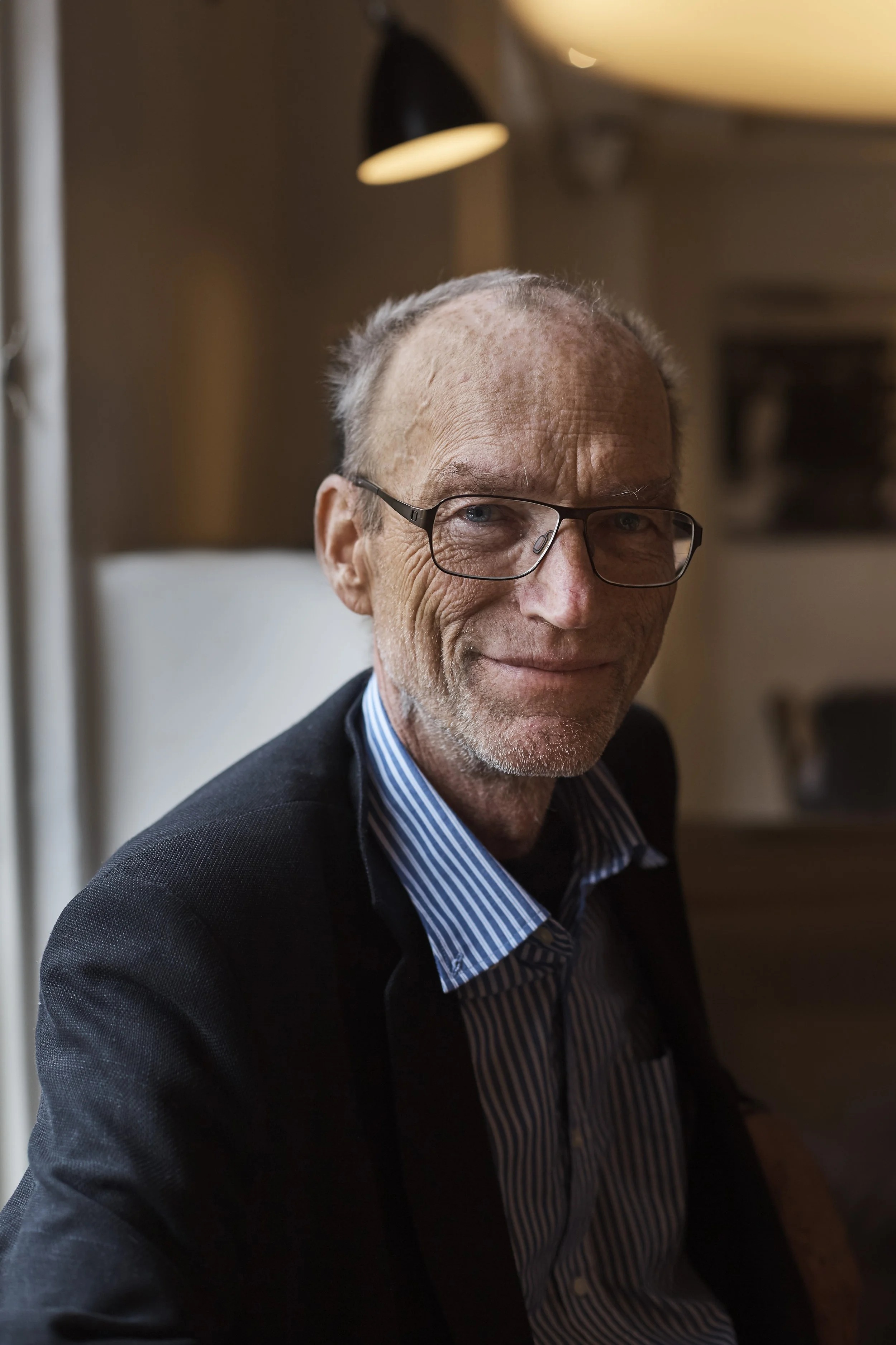 A close-up of an older man with glasses, smiling slightly, wearing a black jacket and a blue-striped shirt, sitting indoors with warm lighting and blurred background.