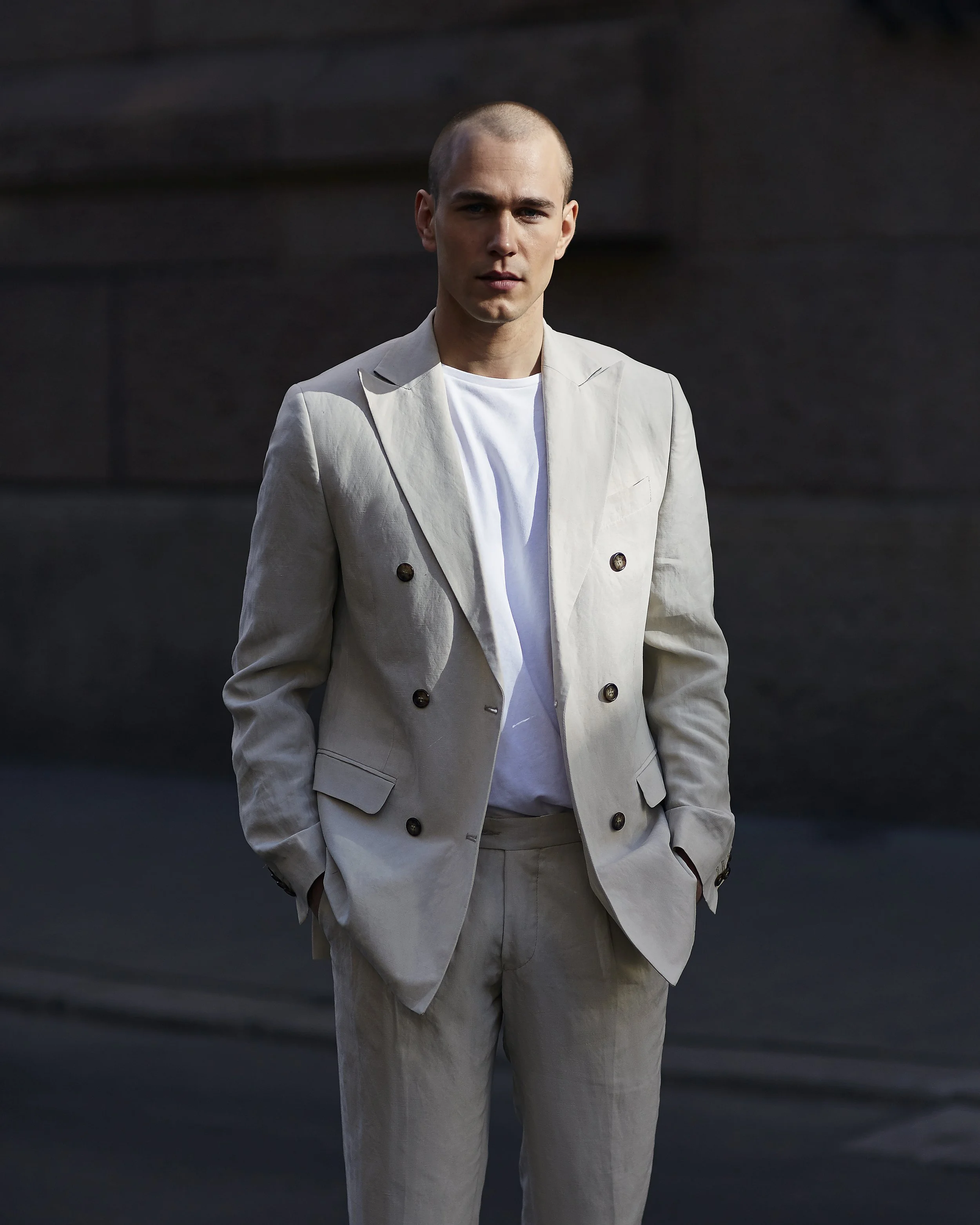 A young man in a light beige suit with a white shirt standing outdoors with hands in pockets, facing the camera.