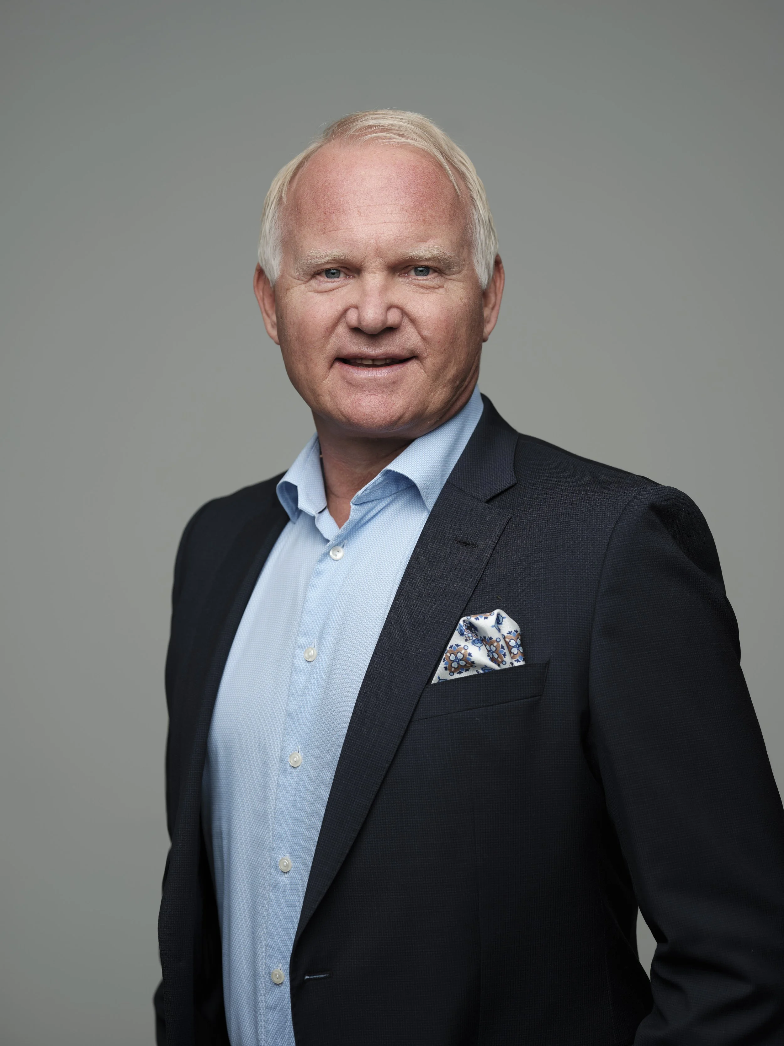 Portrait of a middle-aged man with light hair wearing a navy blazer, light blue shirt, and patterned pocket square, against a neutral gray background.