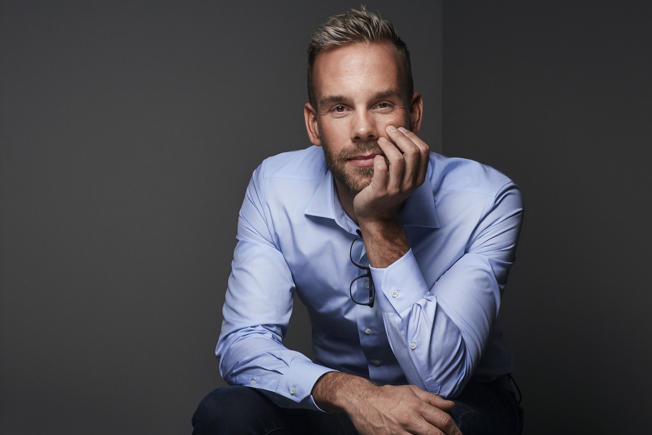 A man with blond hair, beard, wearing a light blue shirt, is resting his head on his hand and looking into the camera against a dark gray background.