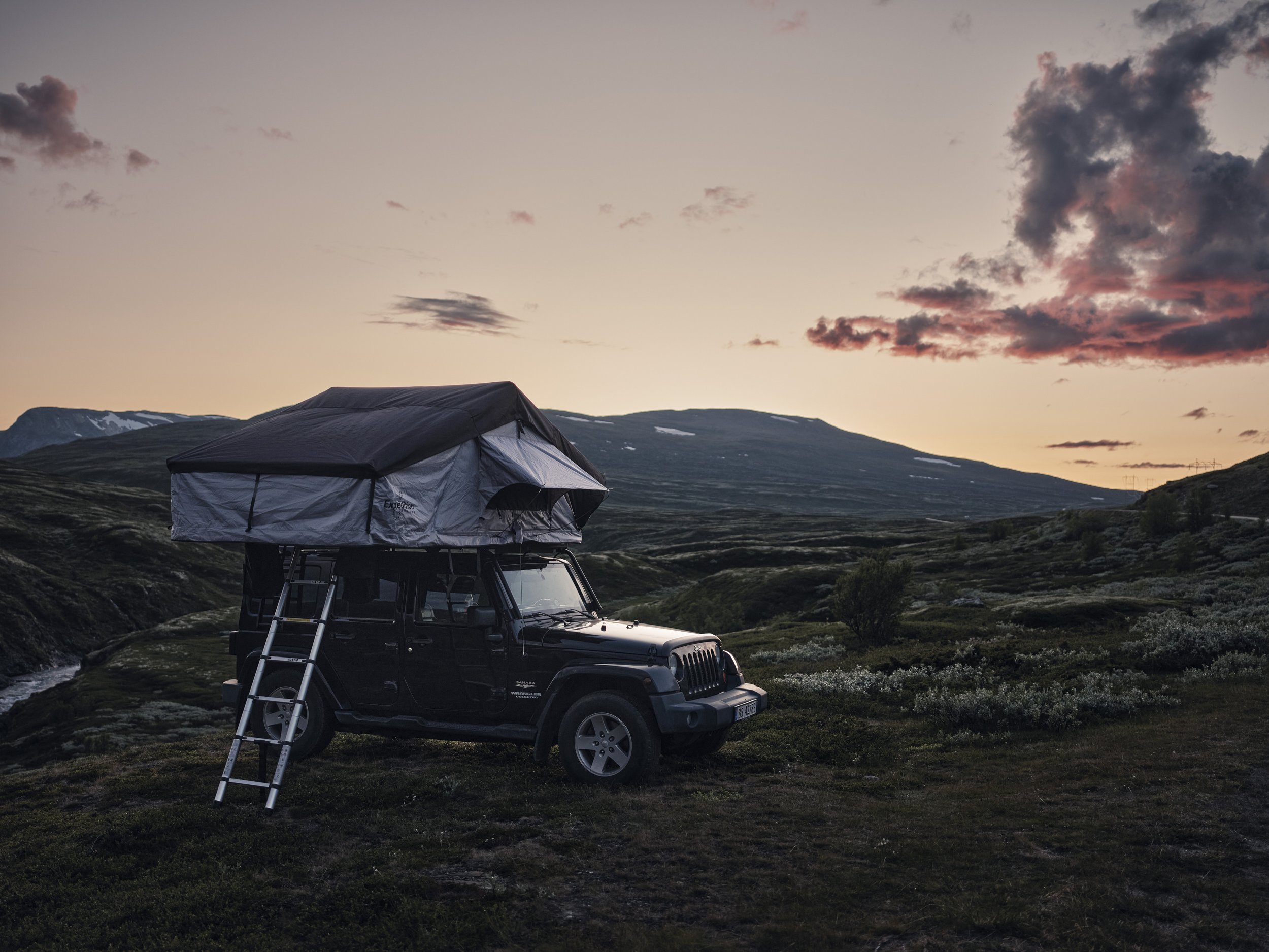 A black Jeep with a rooftop tent and ladder parked on a grassy hillside during sunset with mountains and cloudy sky in the background.