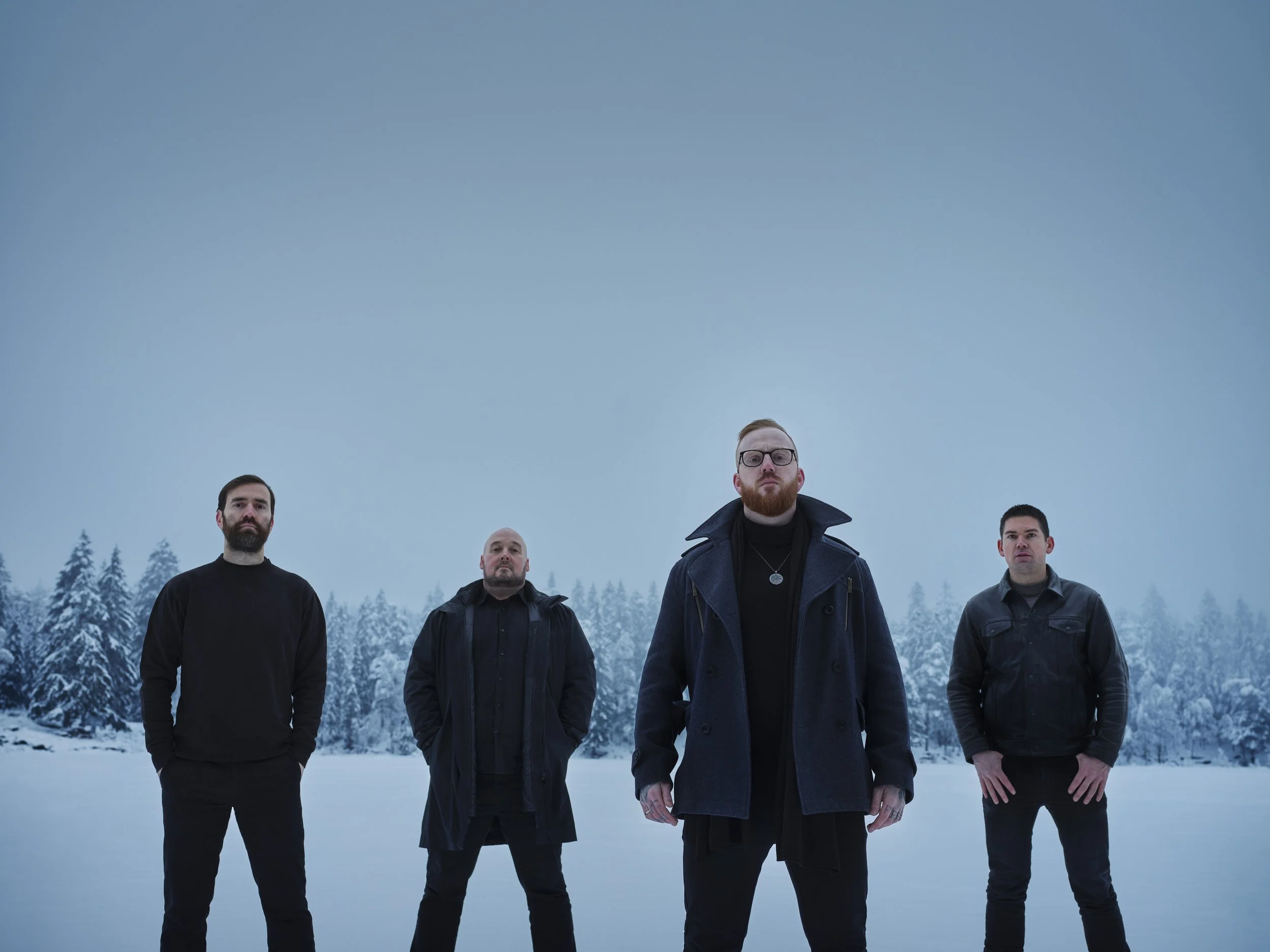 Four men standing outdoors in a snowy landscape with snow-covered trees in the background, facing the camera.