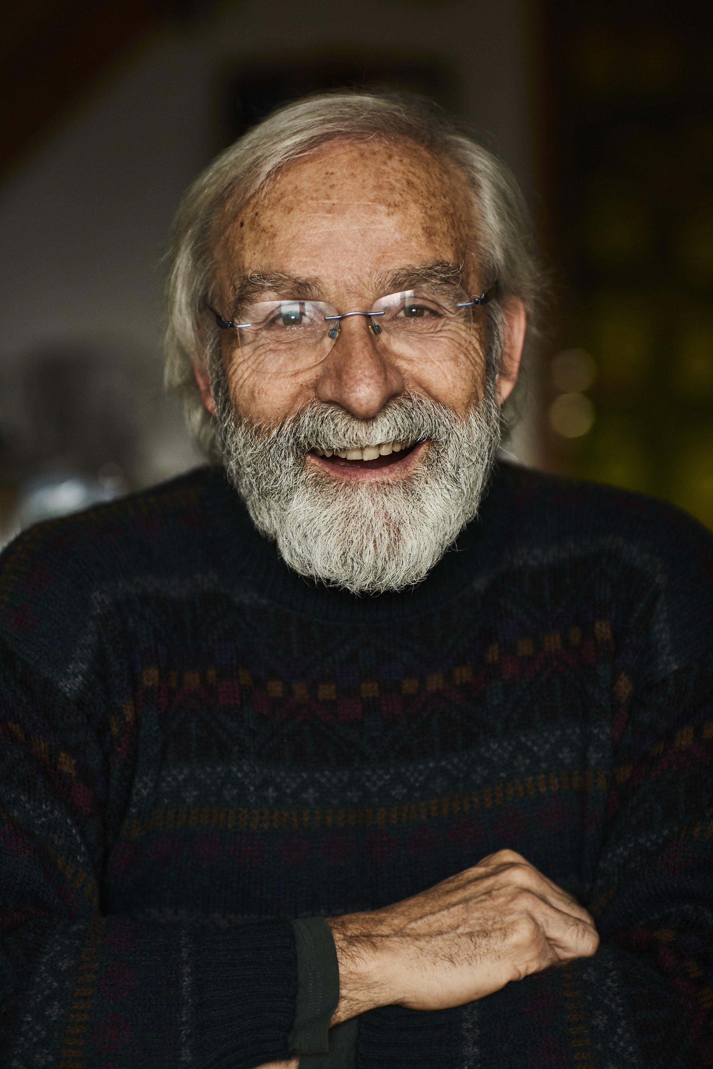 A smiling elderly man with a white beard, glasses, and light skin, wearing a dark patterned sweater, sitting with arms crossed. Portrait. Editorial.