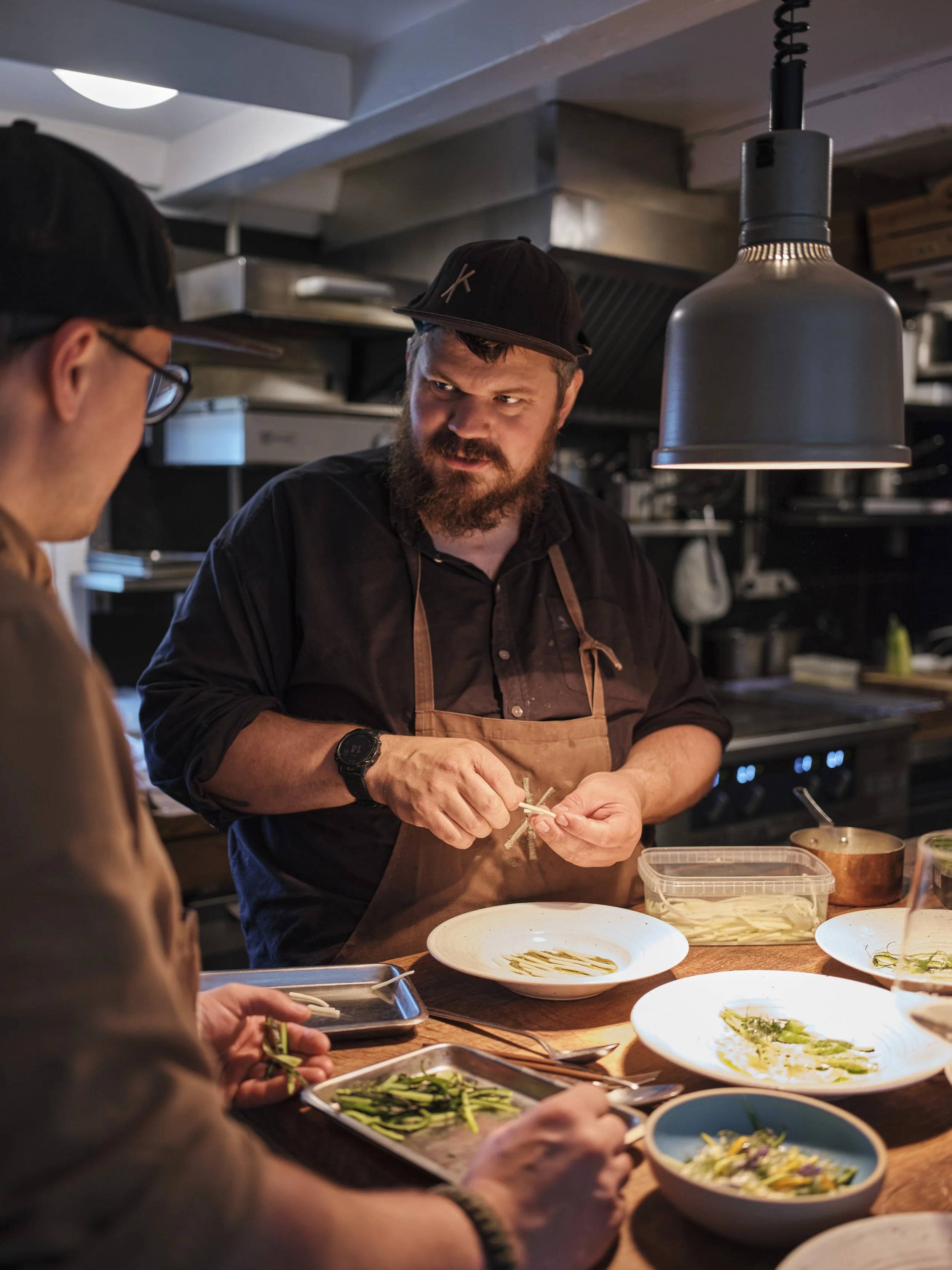 Two men in a restaurant kitchen preparing dishes. One man with a beard is focused on chopping vegetables while the other is concentrating on plating. The setting is warm and professional, with kitchen equipment visible in the background.