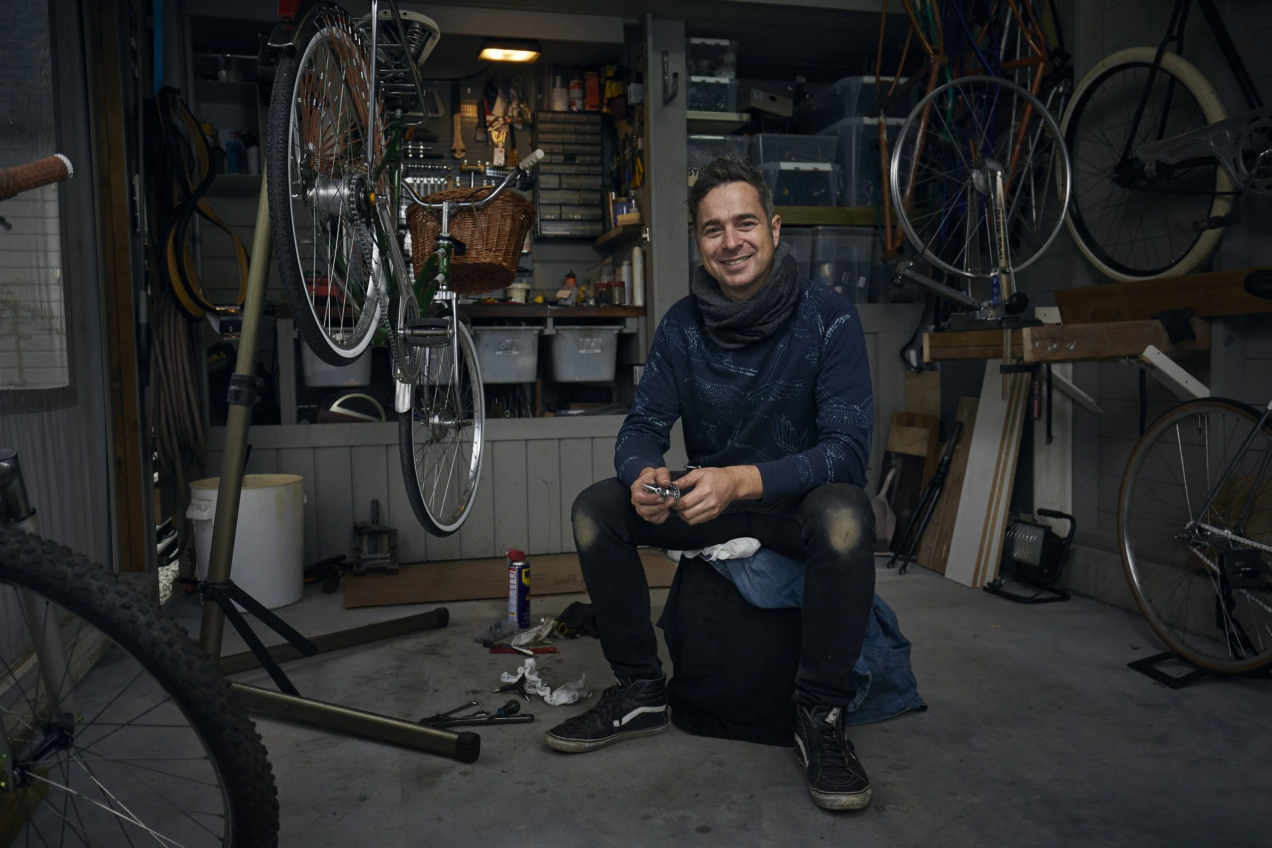 A man sitting on a black bag in a cluttered garage, holding tools and smiling at the camera, surrounded by bicycles and bicycle parts.