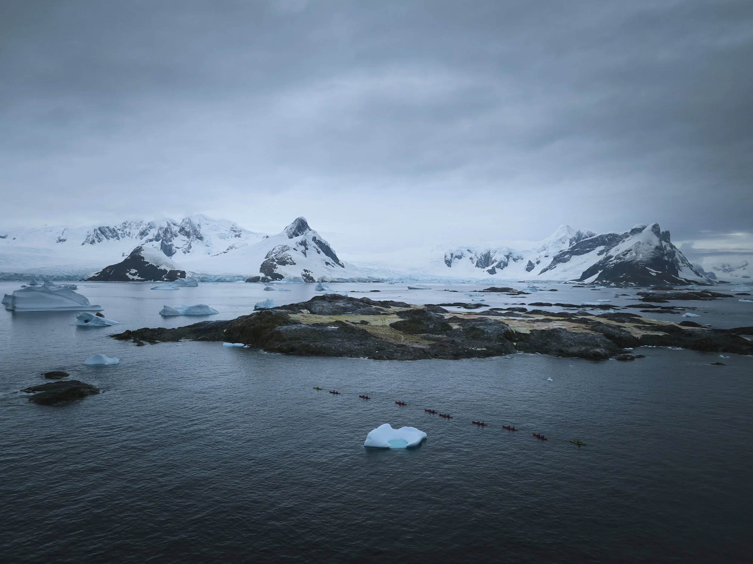 An icy landscape featuring snow-capped mountains, glaciers, and floating icebergs in the water. A line of kayakers paddles through the water near a small iceberg. Antarctica.