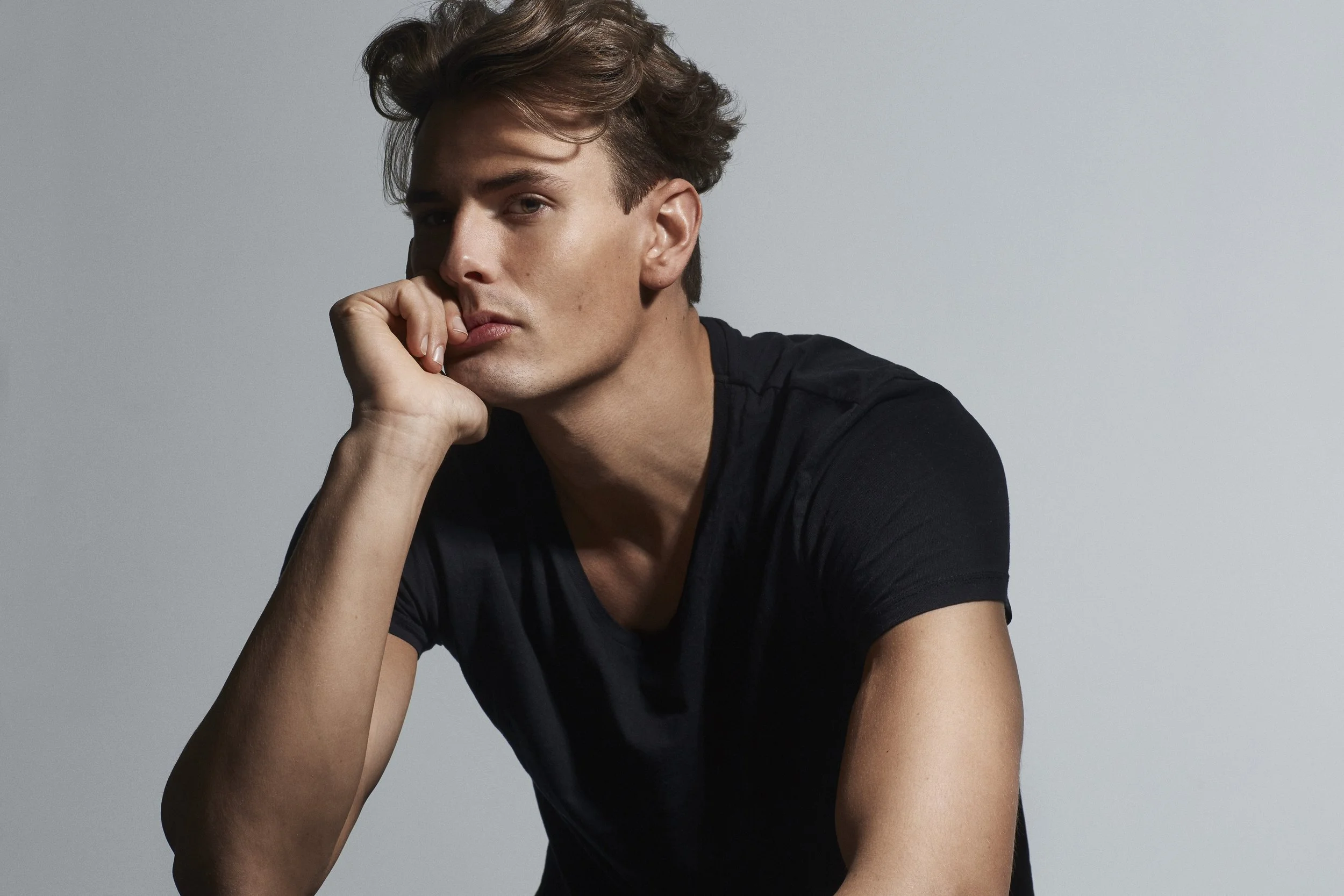 A young man with wavy brown hair, wearing a black t-shirt, resting his chin on his hand against a plain gray background.