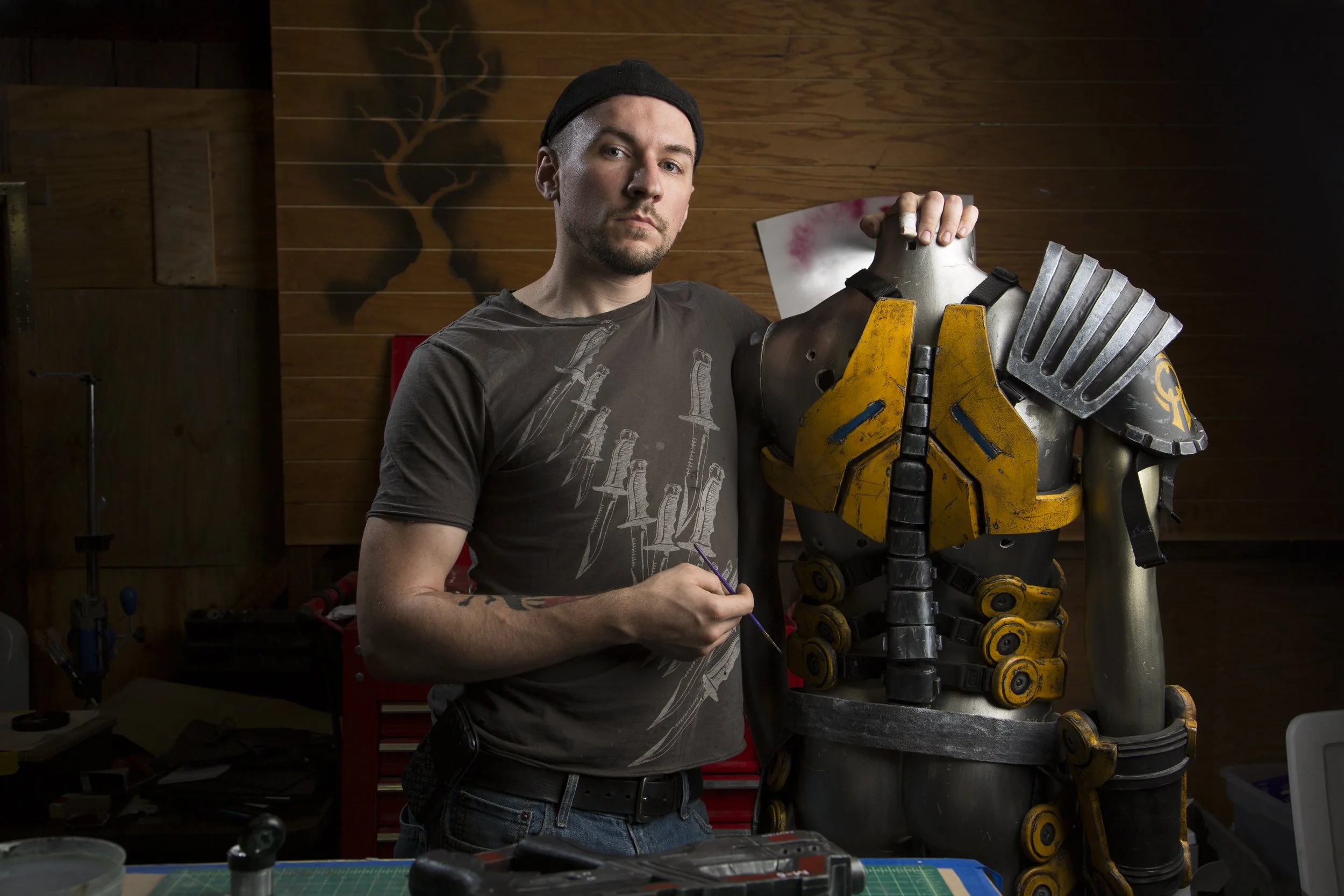 A man standing next to a life-sized robot sculpture, holding a paintbrush in his right hand and wearing a dark t-shirt with knife designs and a black beanie. The robot has a yellow chest plate, metallic shoulder armor, and various mechanical details.