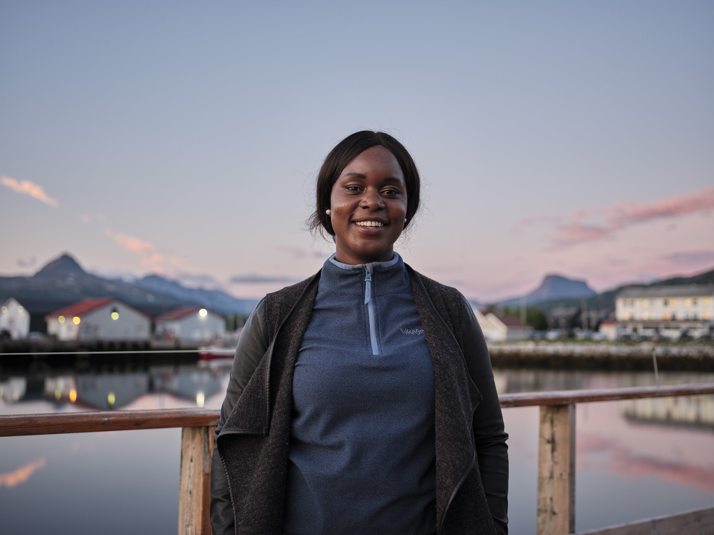 A woman smiling outdoors near a waterfront with mountains in the background at sunset.