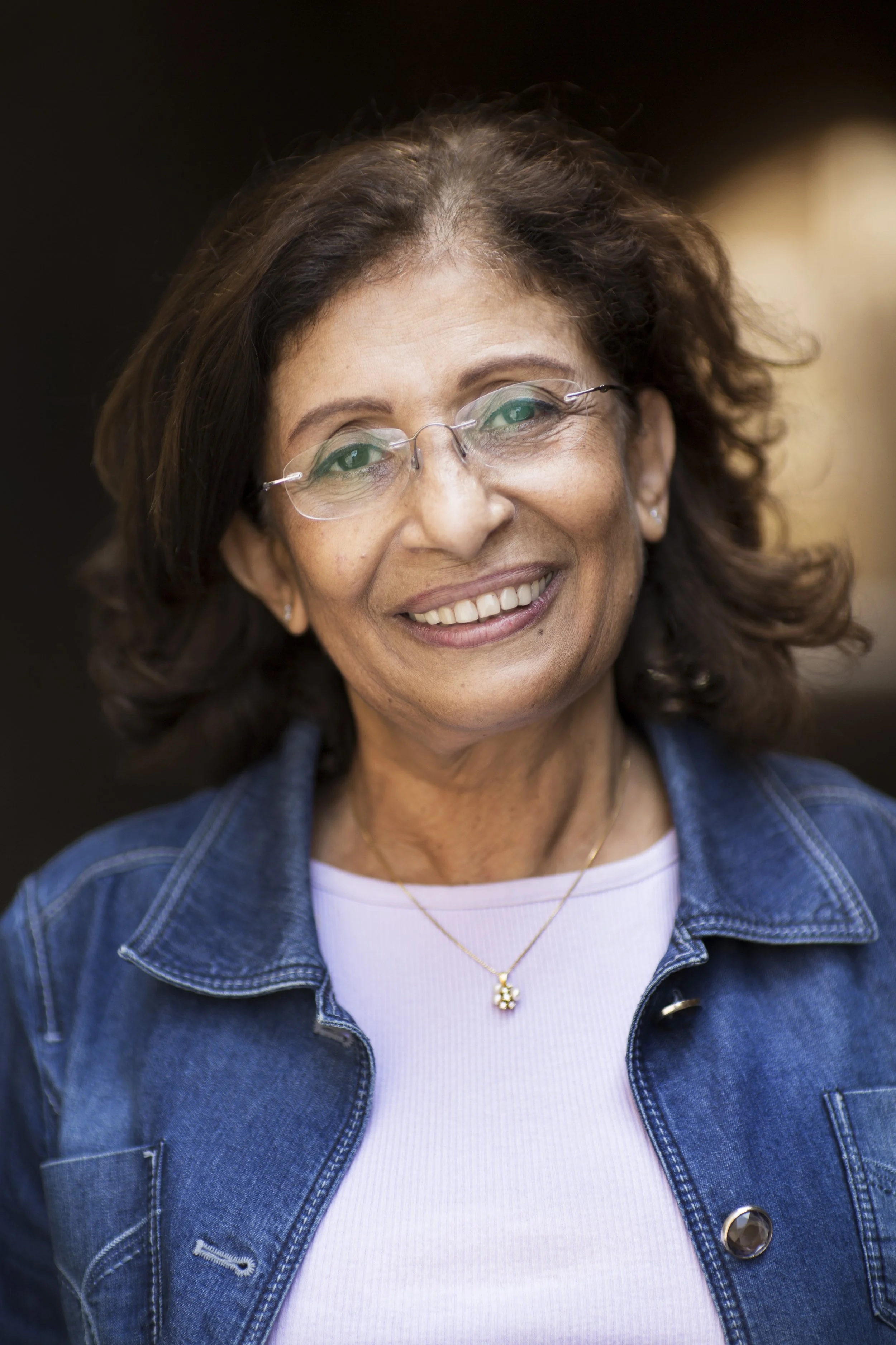 Close-up of a smiling woman with glasses, wearing a denim jacket and a white top, against a dark background.