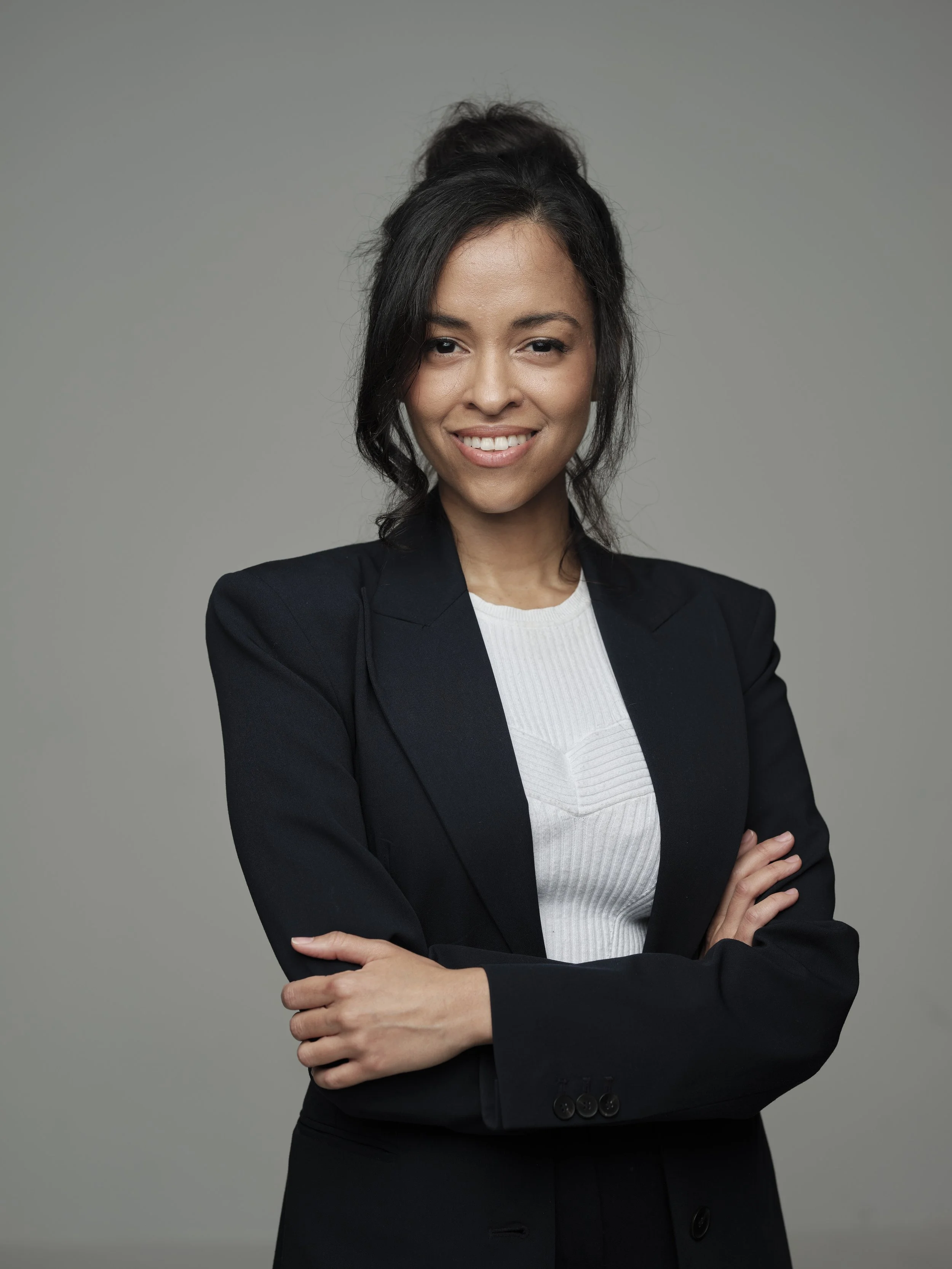 Businesswoman with crossed arms, smiling, and wearing a black blazer and white top against a plain gray background.