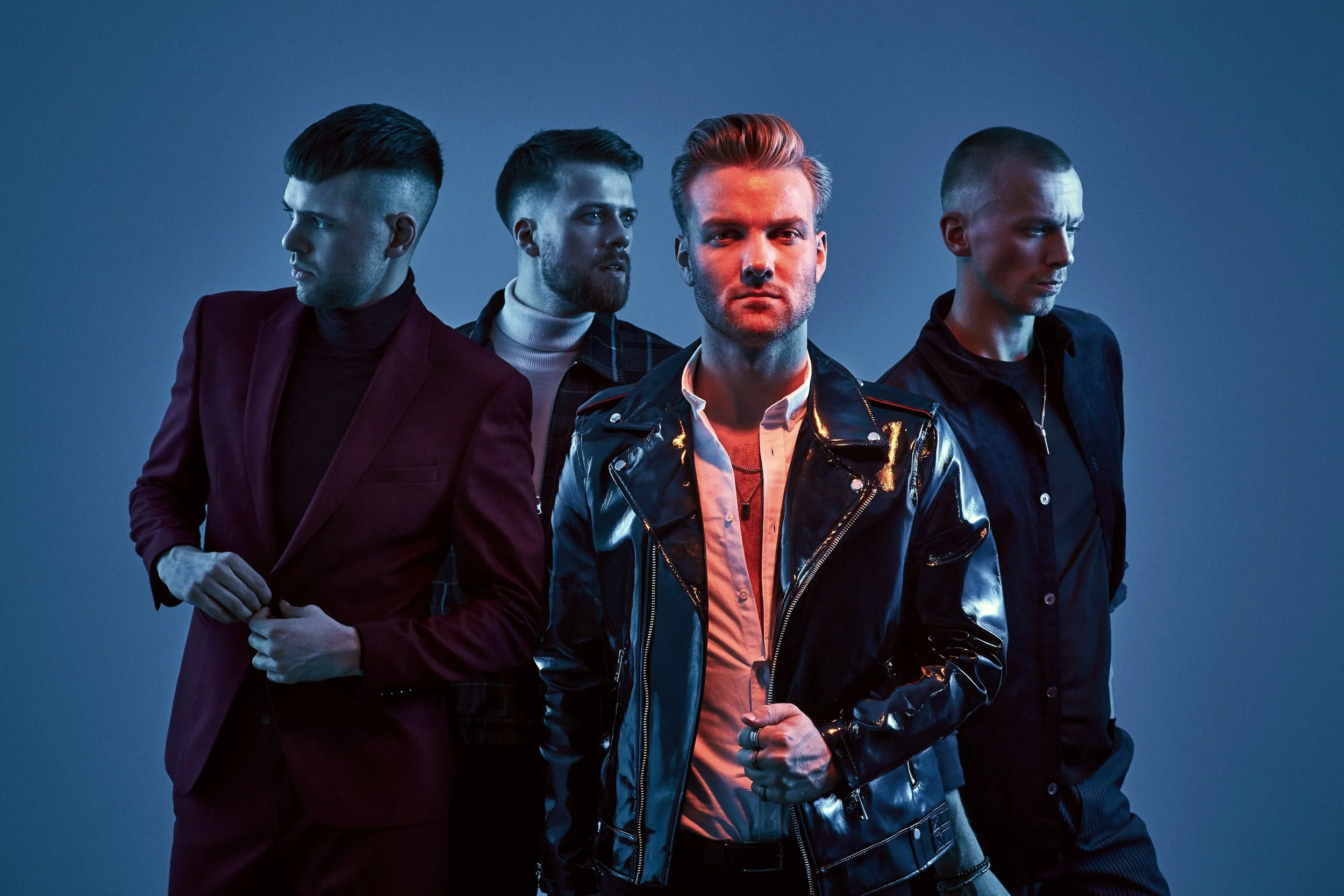 Four men standing together against a blue background, with the front man wearing a shiny black leather jacket and the others dressed in darker clothing.