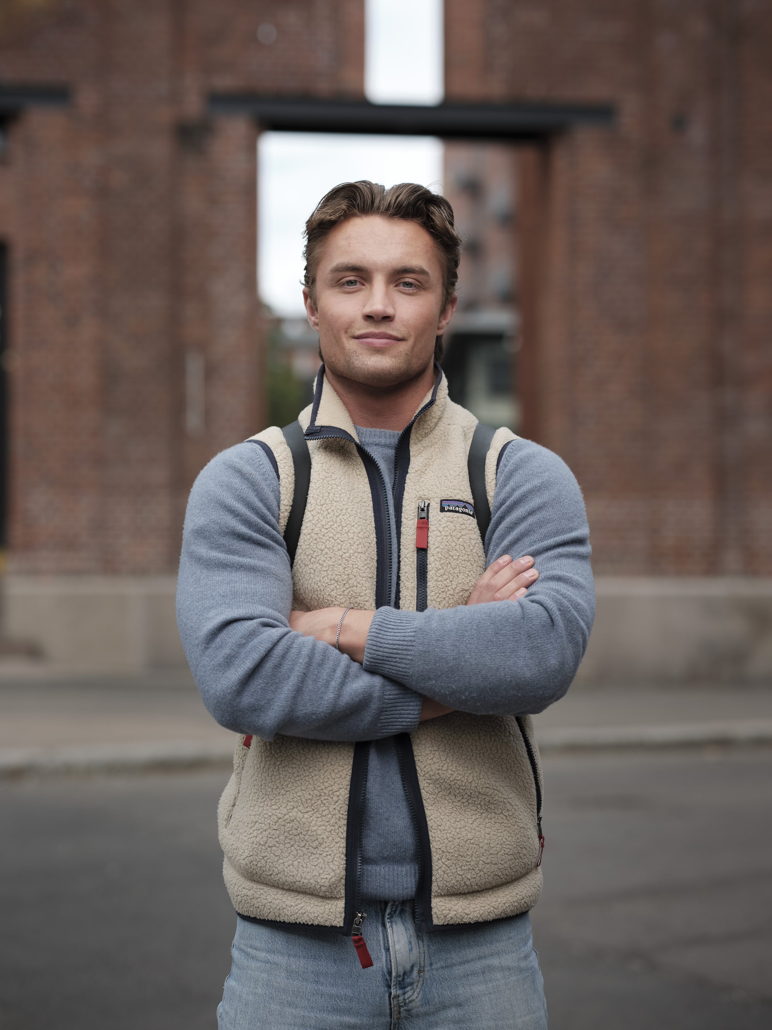 A young man with crossed arms standing outdoors in front of a brick building, wearing a Patagonia fleece vest and backpack.