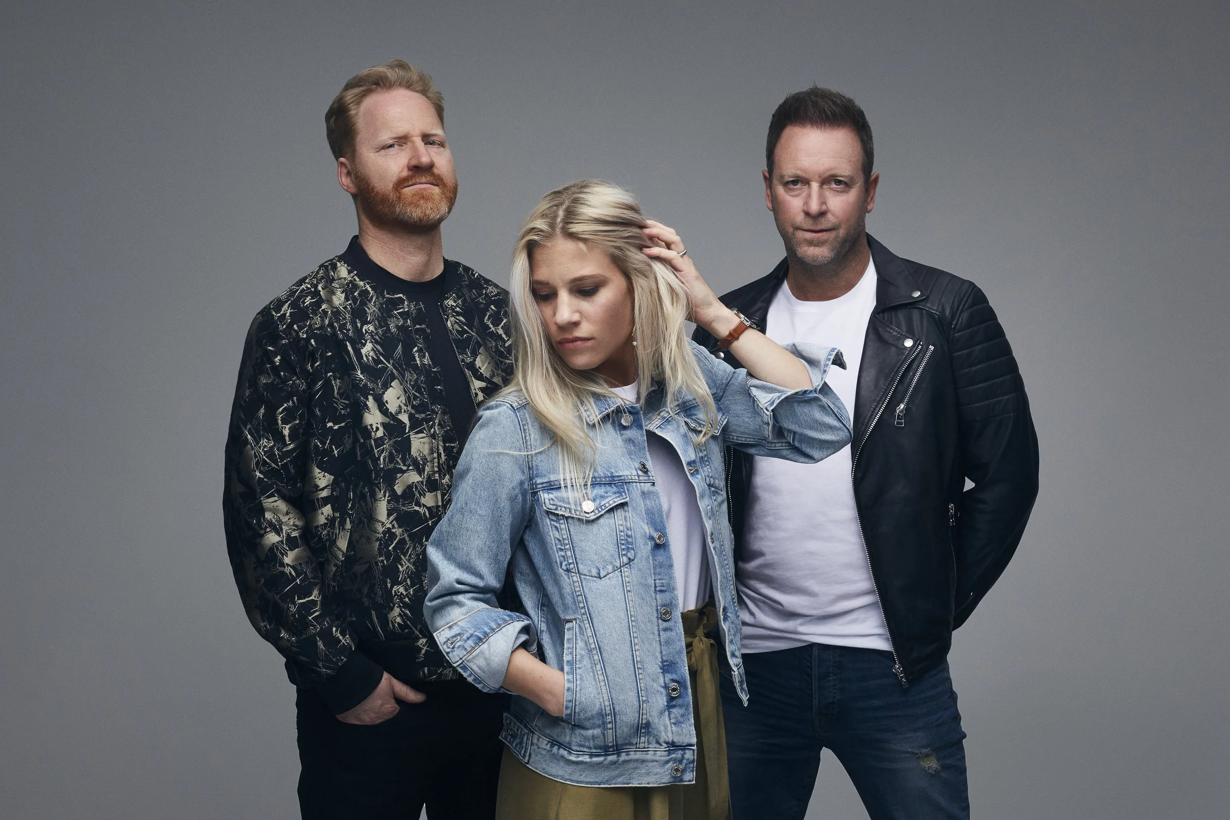 Three people standing together against a gray background, a woman in front wearing a denim jacket looking down, a man with sunglasses and a patterned jacket on the left, and a man in a leather jacket on the right.
