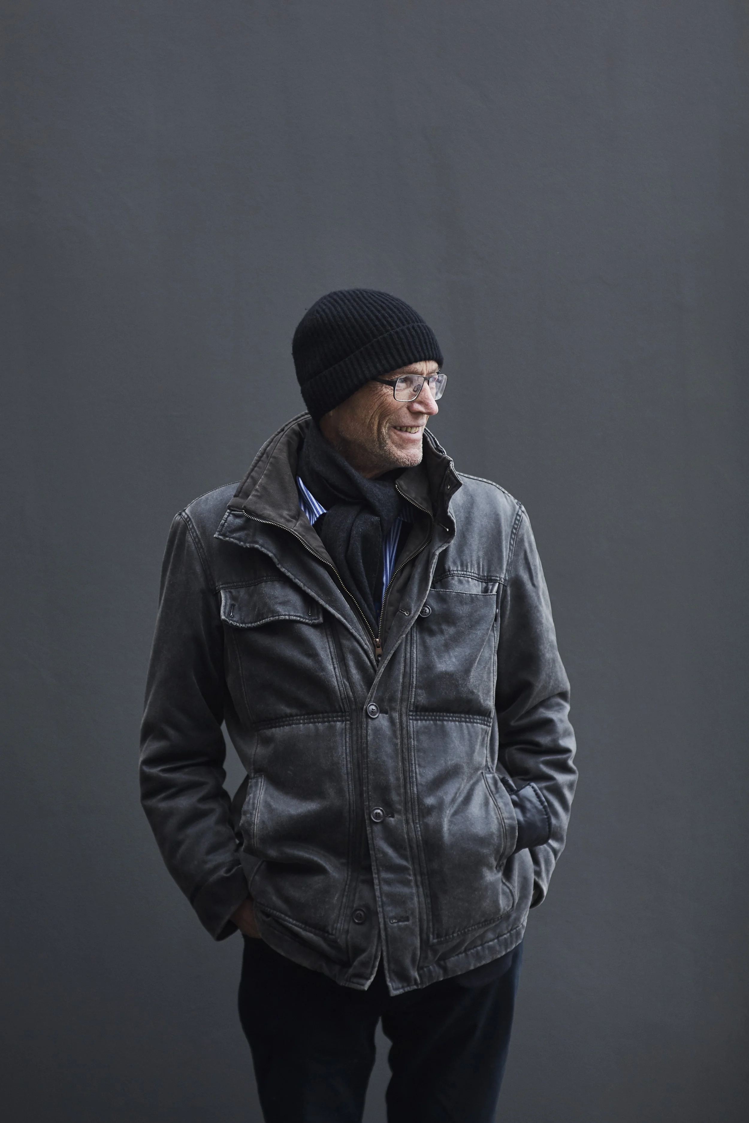 A man wearing a black knit beanie, glasses, a dark gray jacket, and a black scarf stands against a plain gray background, smiling and looking to his left.
