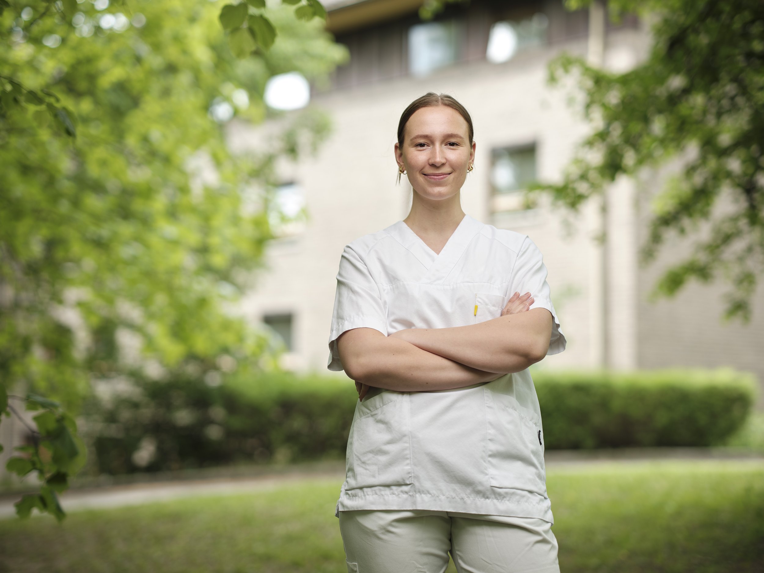 A young woman in white nurse scrubs standing outdoors with arms crossed, smiling, with a residential building and trees in the background.