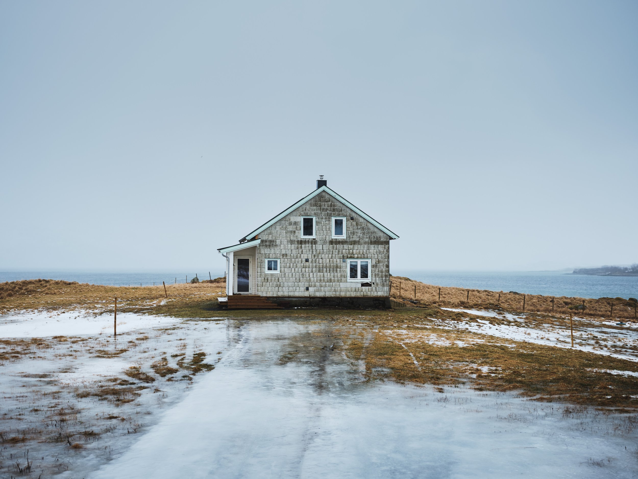 A house with a gray shingle exterior and white trim, located outdoors near water, with a dirt pathway in front, some patches of snow or ice, and a cloudy sky in the background.