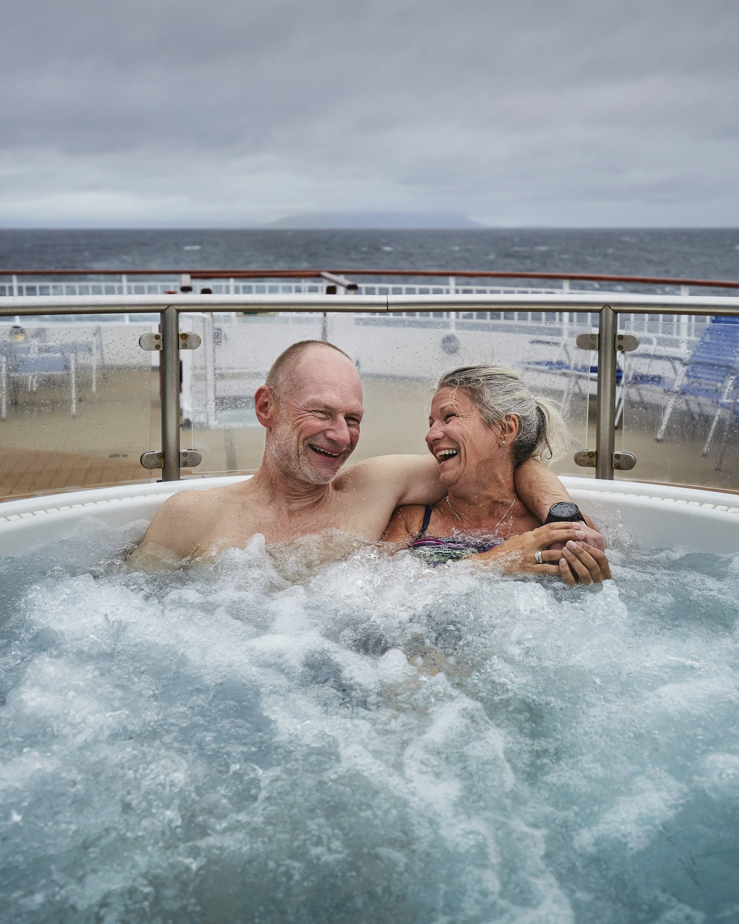 An elderly couple enjoying a hot tub on a cruise ship in rough weather, with the ocean and gray sky in the background.