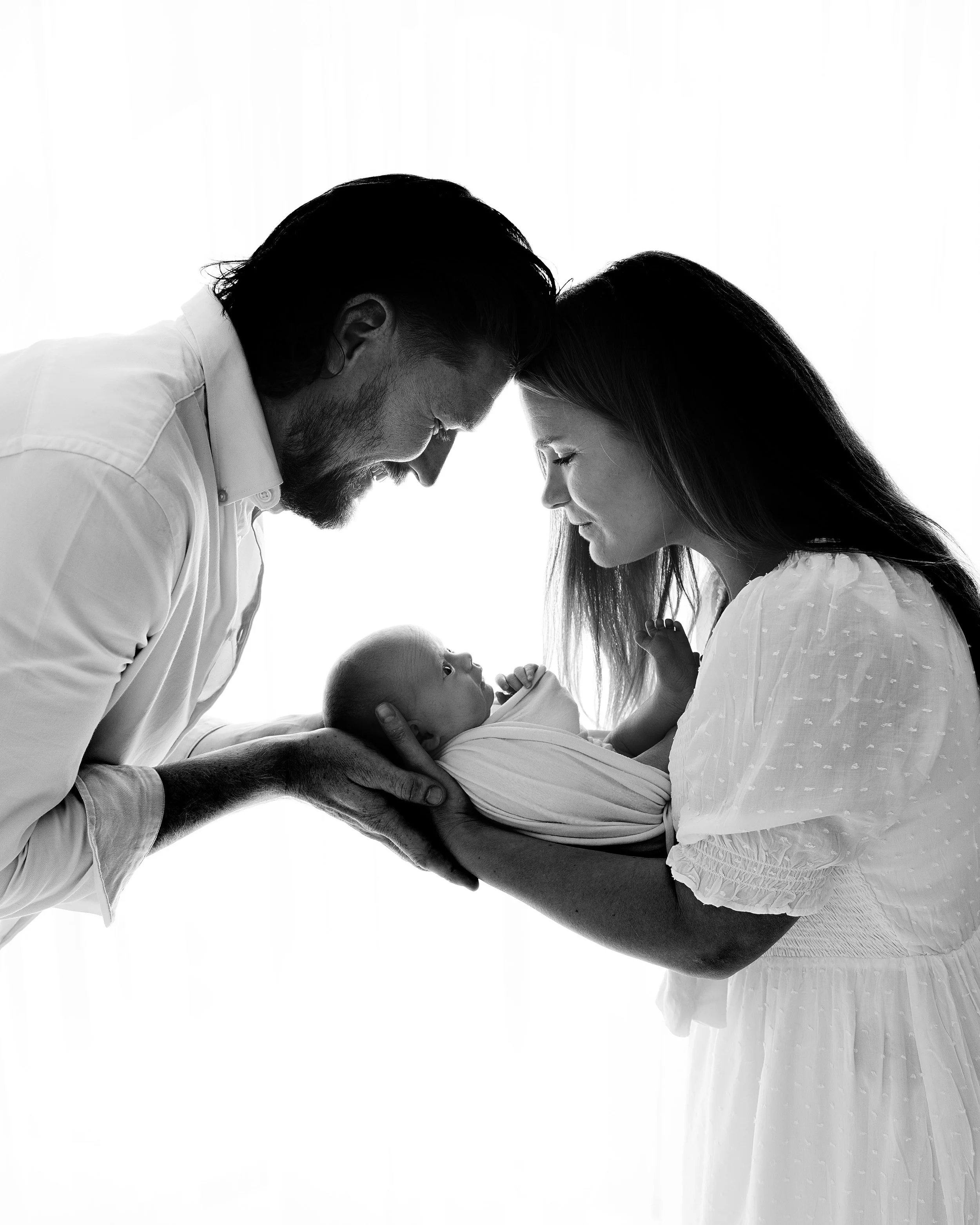 A black-and-white photo of a family with a newborn baby, the father and mother touching foreheads and looking at the baby.