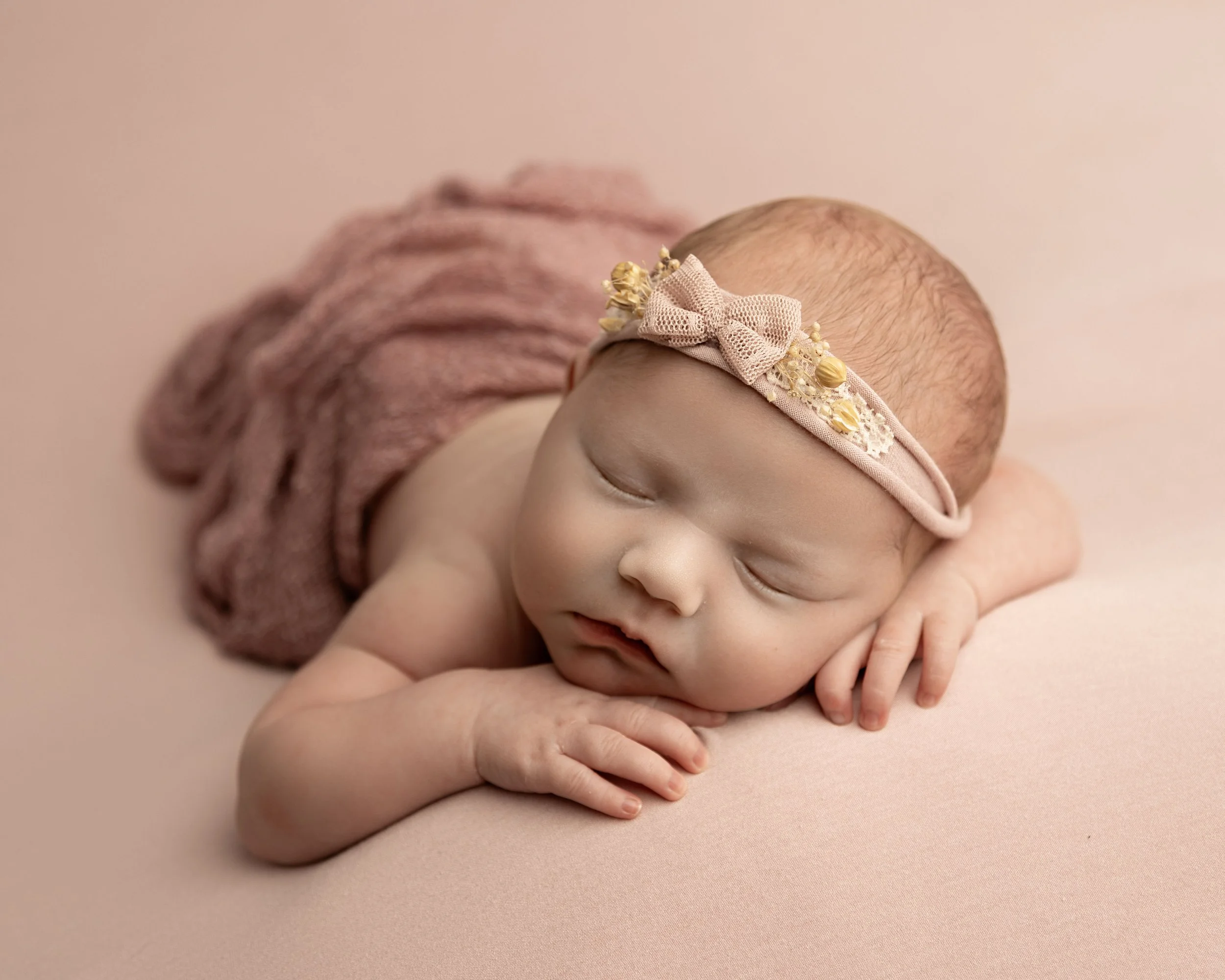 A sleeping newborn baby with a pink headband and a pink knitted wrap on a soft pink background.
