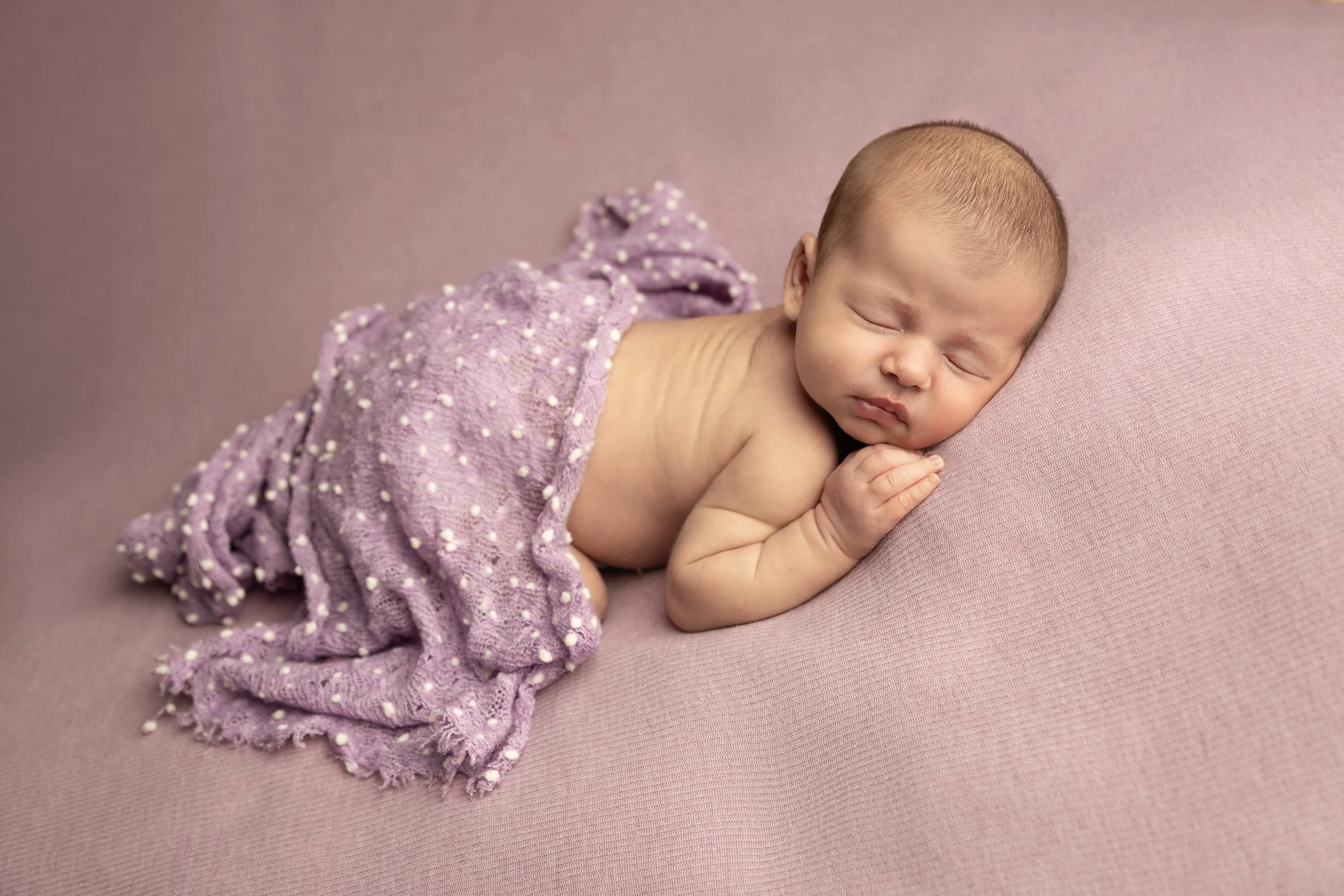 A sleeping newborn baby lying on a pink surface, wrapped in a purple knitted blanket with white dots, with one hand touching its cheek.