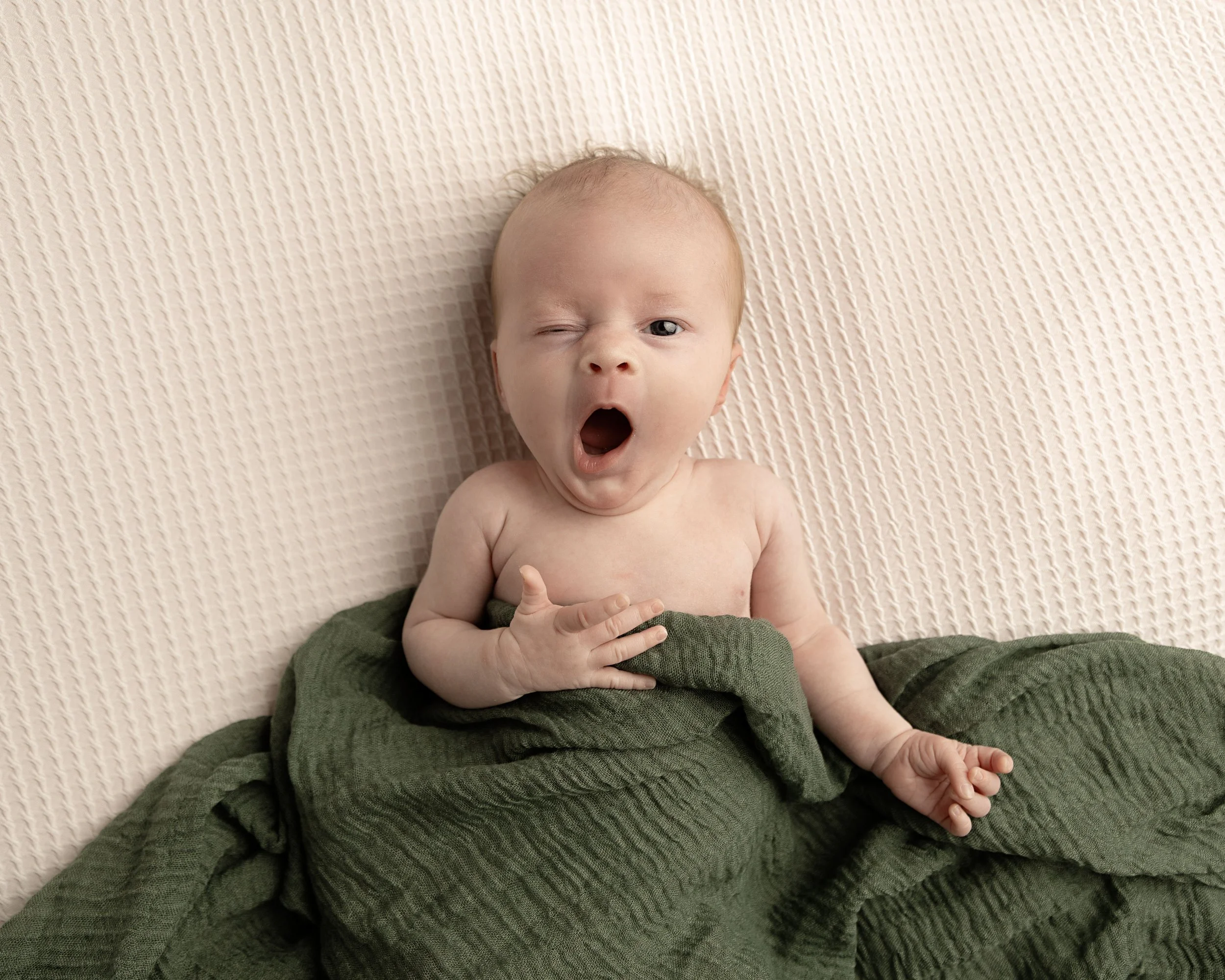 A baby with light skin and sparse hair lying on a textured beige surface, partially covered with a green blanket, yawning with one eye closed.