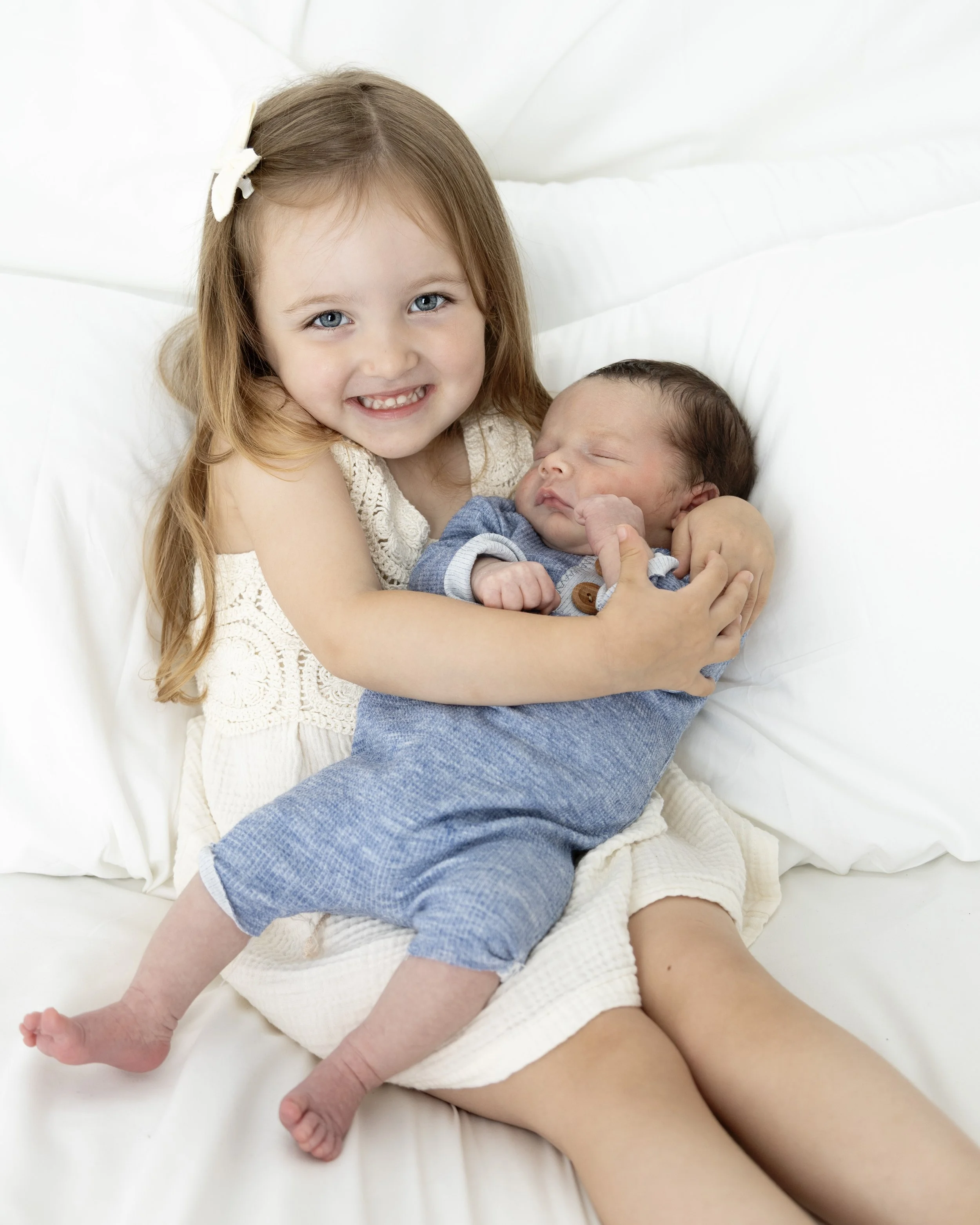 A young girl with long red hair, blue eyes, and a white bow, smiling while holding a sleeping newborn baby with dark hair dressed in blue, on a bed with white sheets.