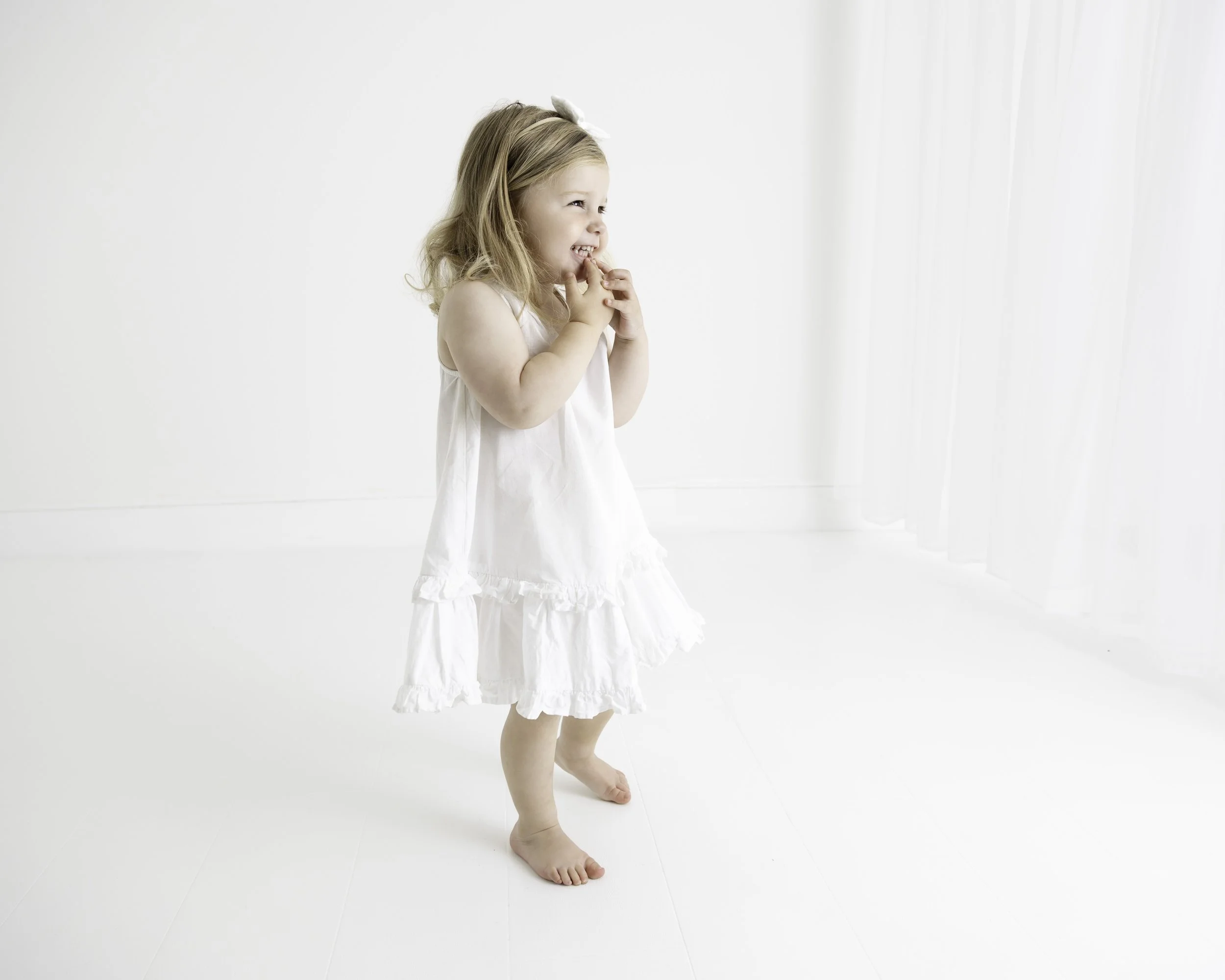 A young girl in a white dress laughing and holding her hands near her face in a bright room with white walls and sheer curtains.