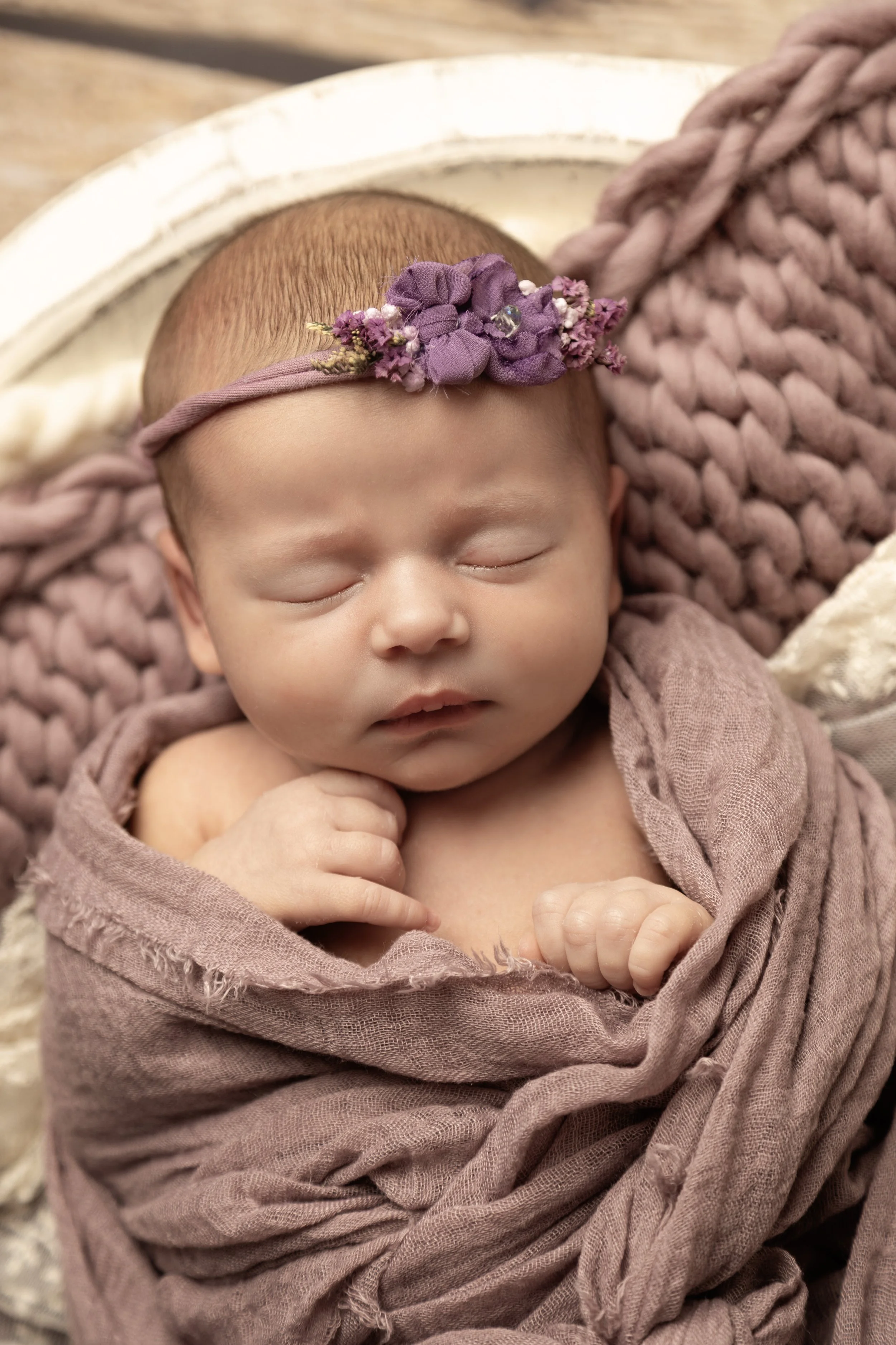 A sleeping baby girl with a purple floral headband, wrapped in a soft pink blanket and resting on a chunky knit pillow.