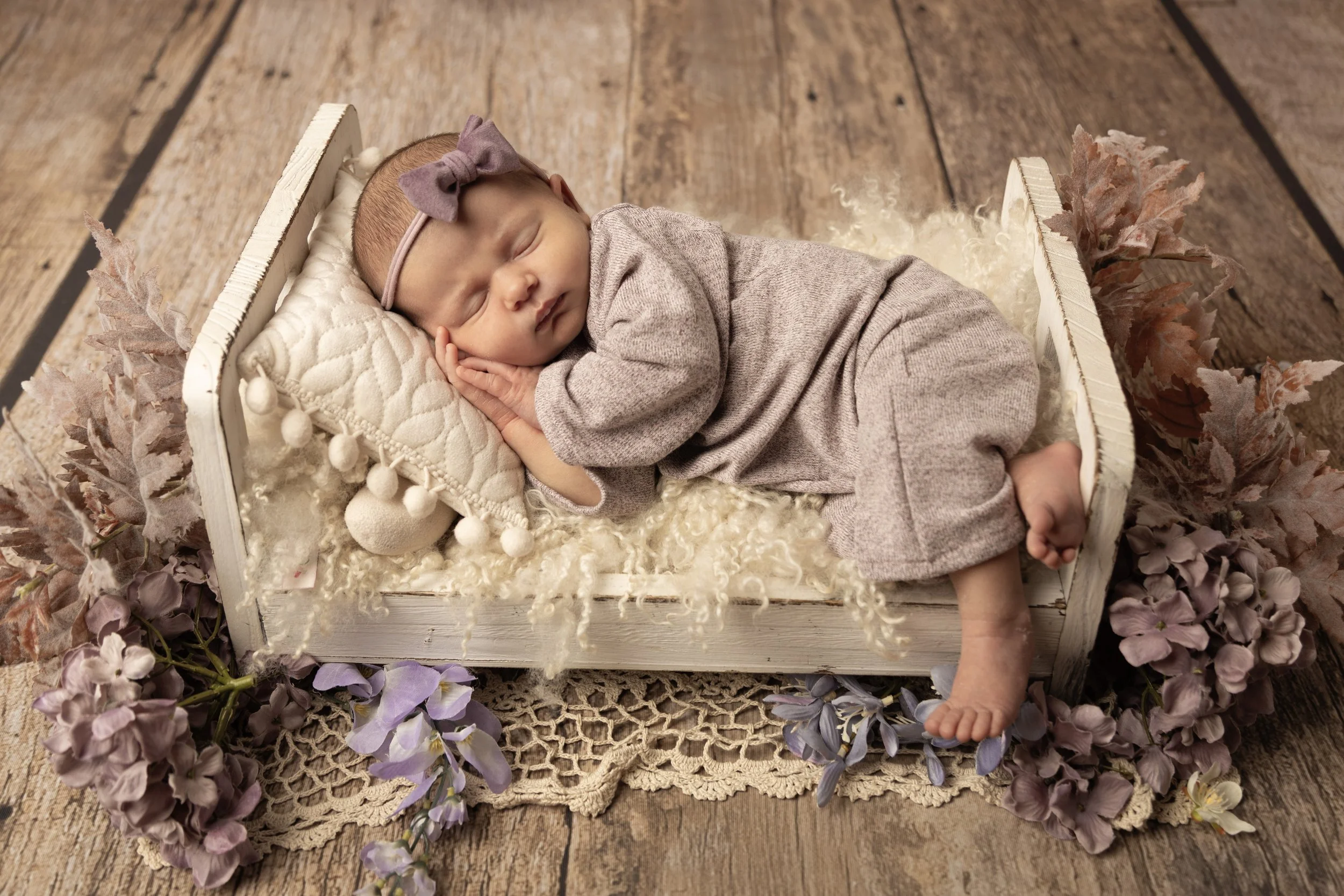 A sleeping baby girl lying in a small white wooden bed with a quilted pillow, on a fluffy white blanket, surrounded by purple and lavender flowers on a wooden floor.