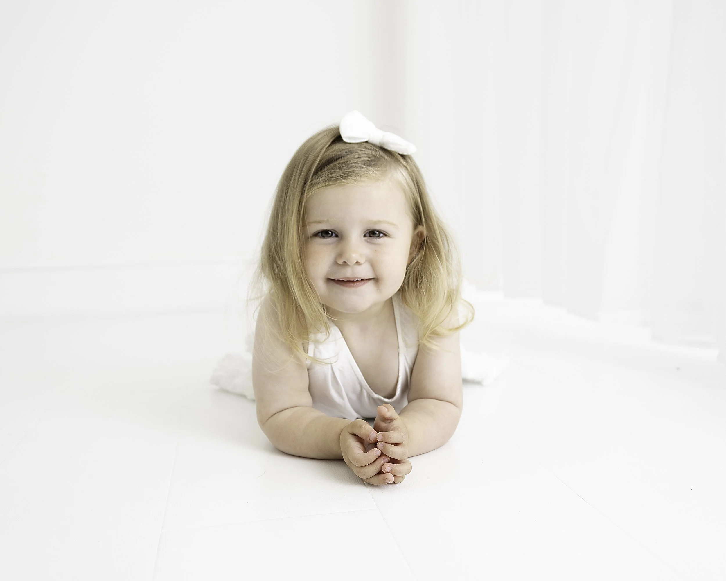A smiling young girl with blonde hair, wearing a white sleeveless top and a white bow in her hair, lying on her stomach on a white floor in a bright, minimalist room with white curtains.