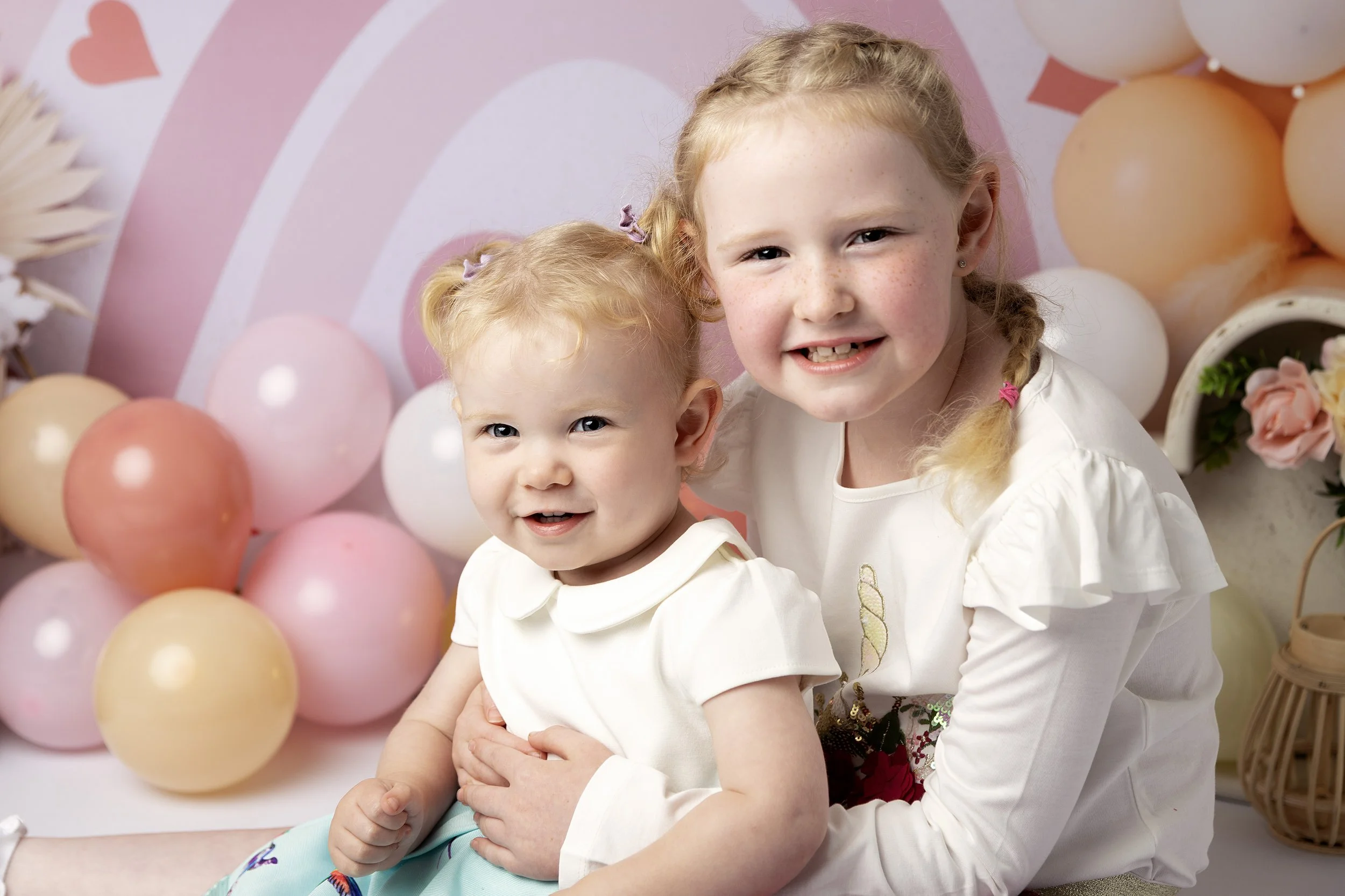 Two young girls, one older and one younger, smiling at the camera with pink, peach, and white balloons in the background.