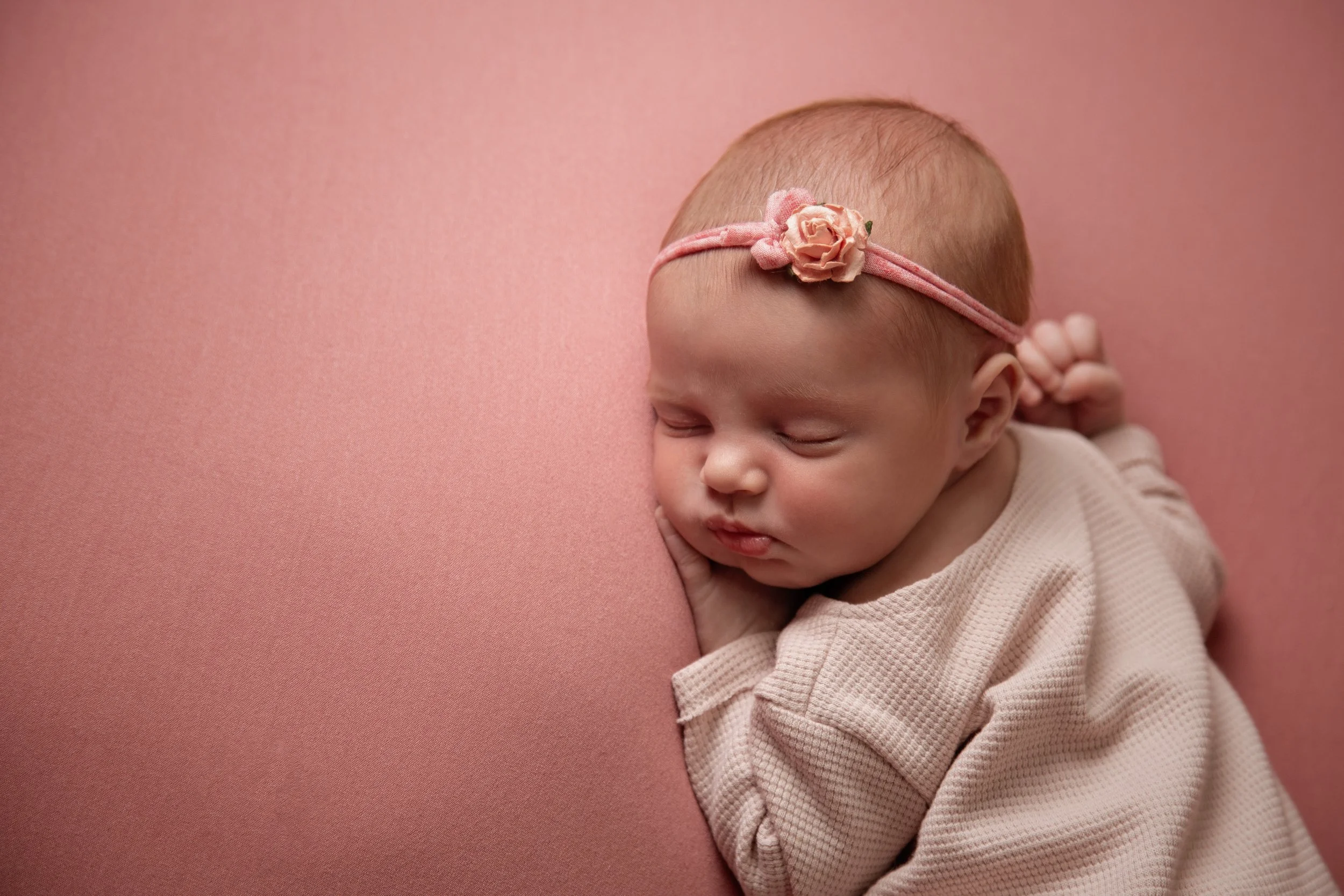 A sleeping baby girl with a pink flower headband, resting on a pink surface.
