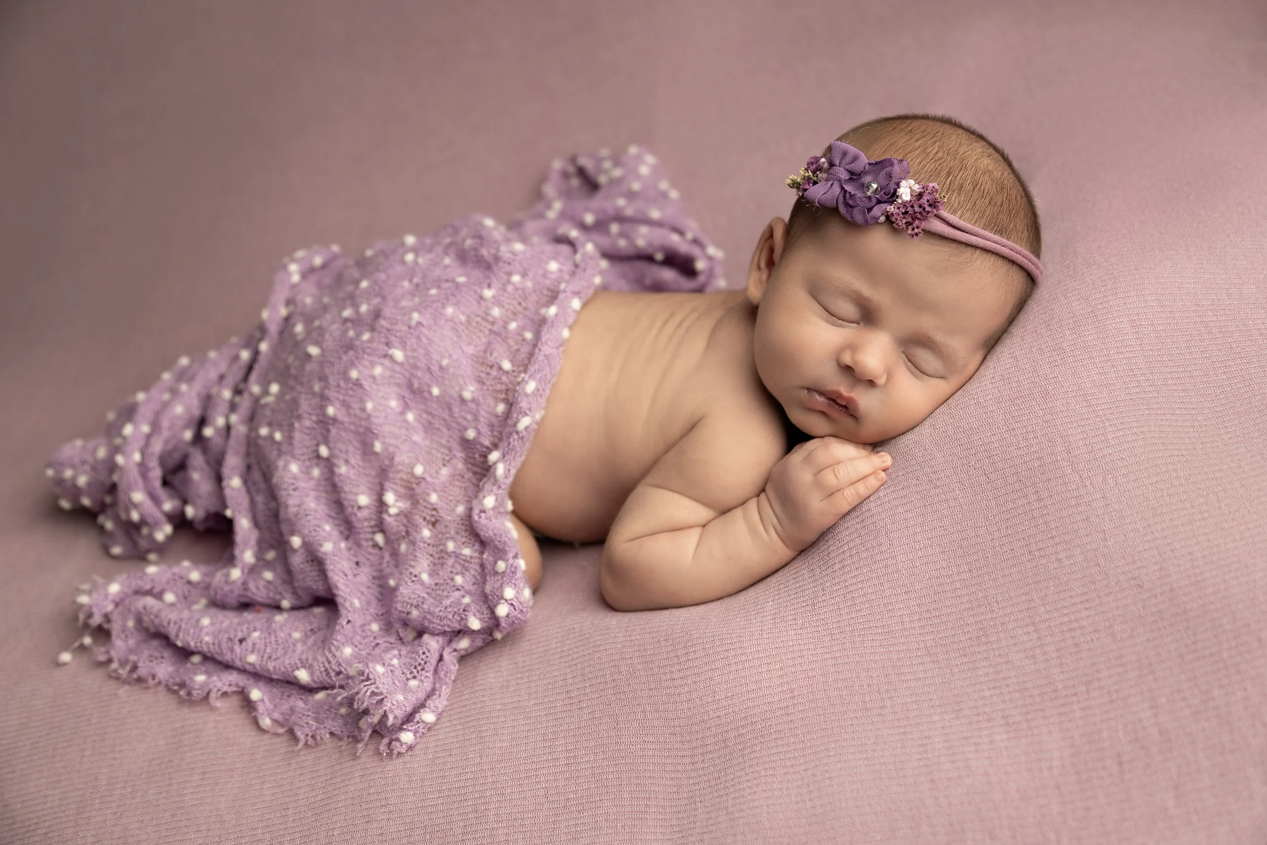 A sleeping baby girl lying on a pink surface, wrapped in a purple knitted blanket with white pom-pom details, wearing a purple headband with purple and white flowers.