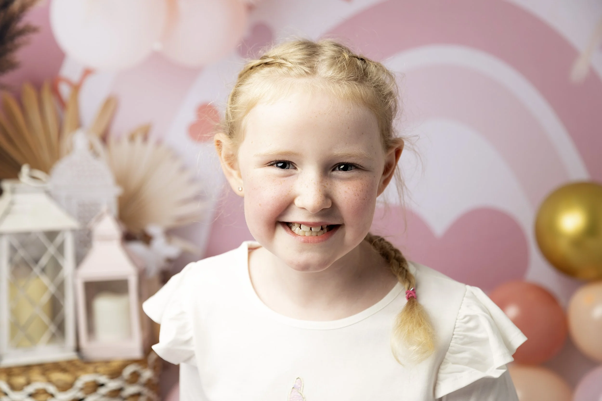 A young girl with blonde hair in a braid, smiling at a birthday celebration with pink and gold balloons in the background.