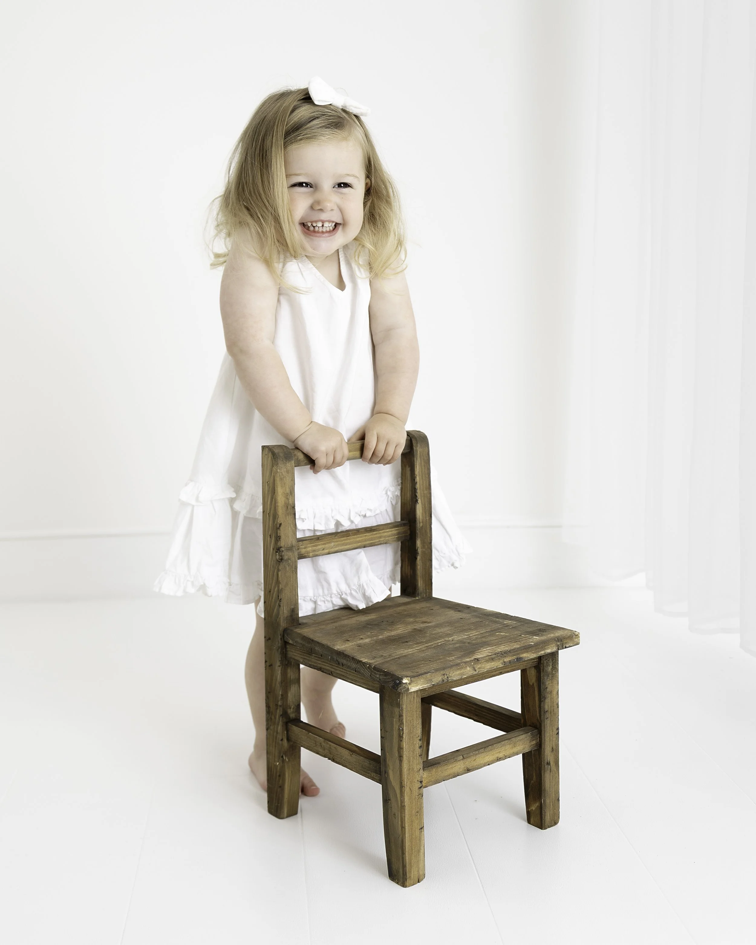 A young girl with blonde hair in a white dress and bow is smiling while holding onto a small wooden chair.
