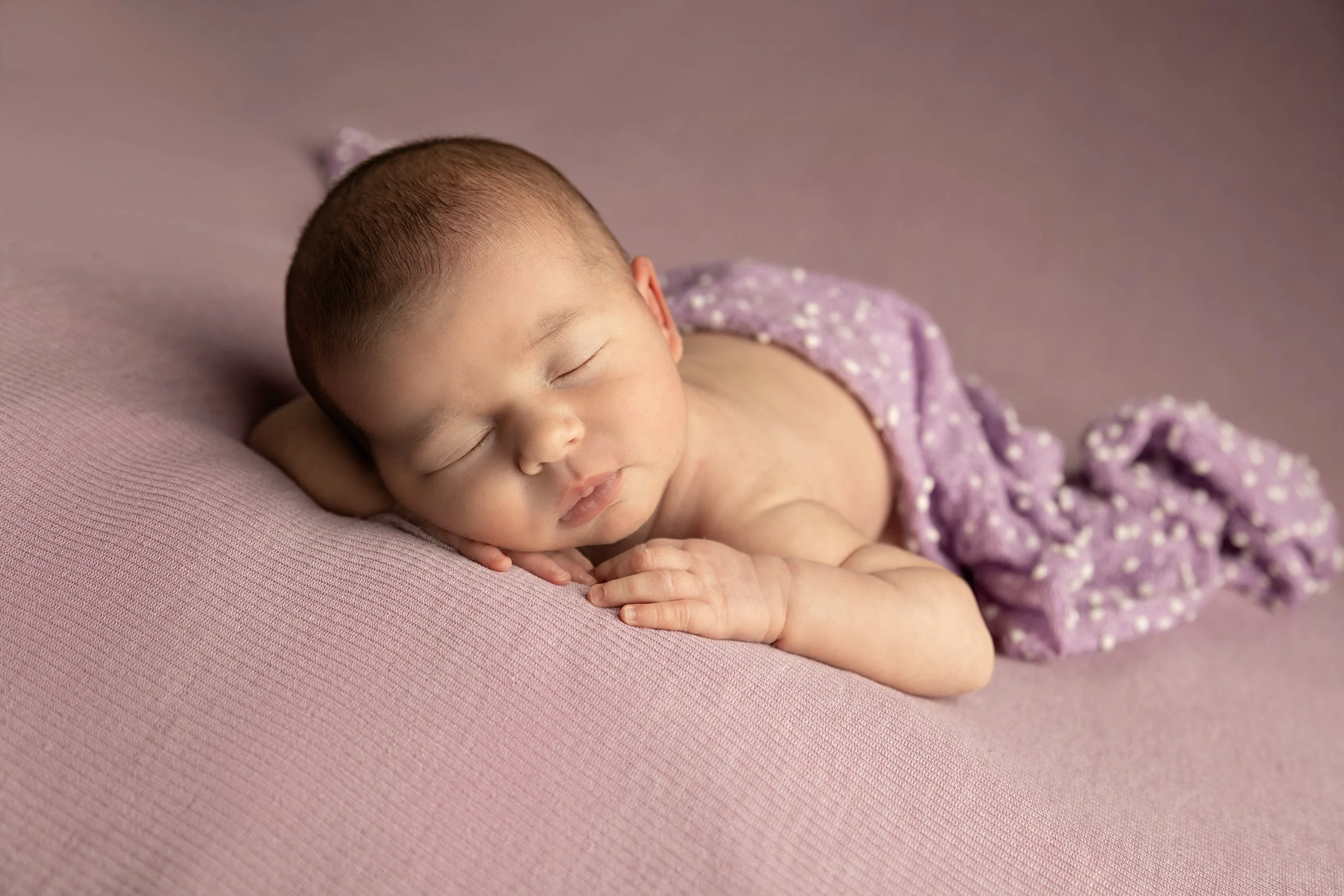 A sleeping baby lying on a pink cushioned surface, wrapped in a purple polka-dotted blanket.