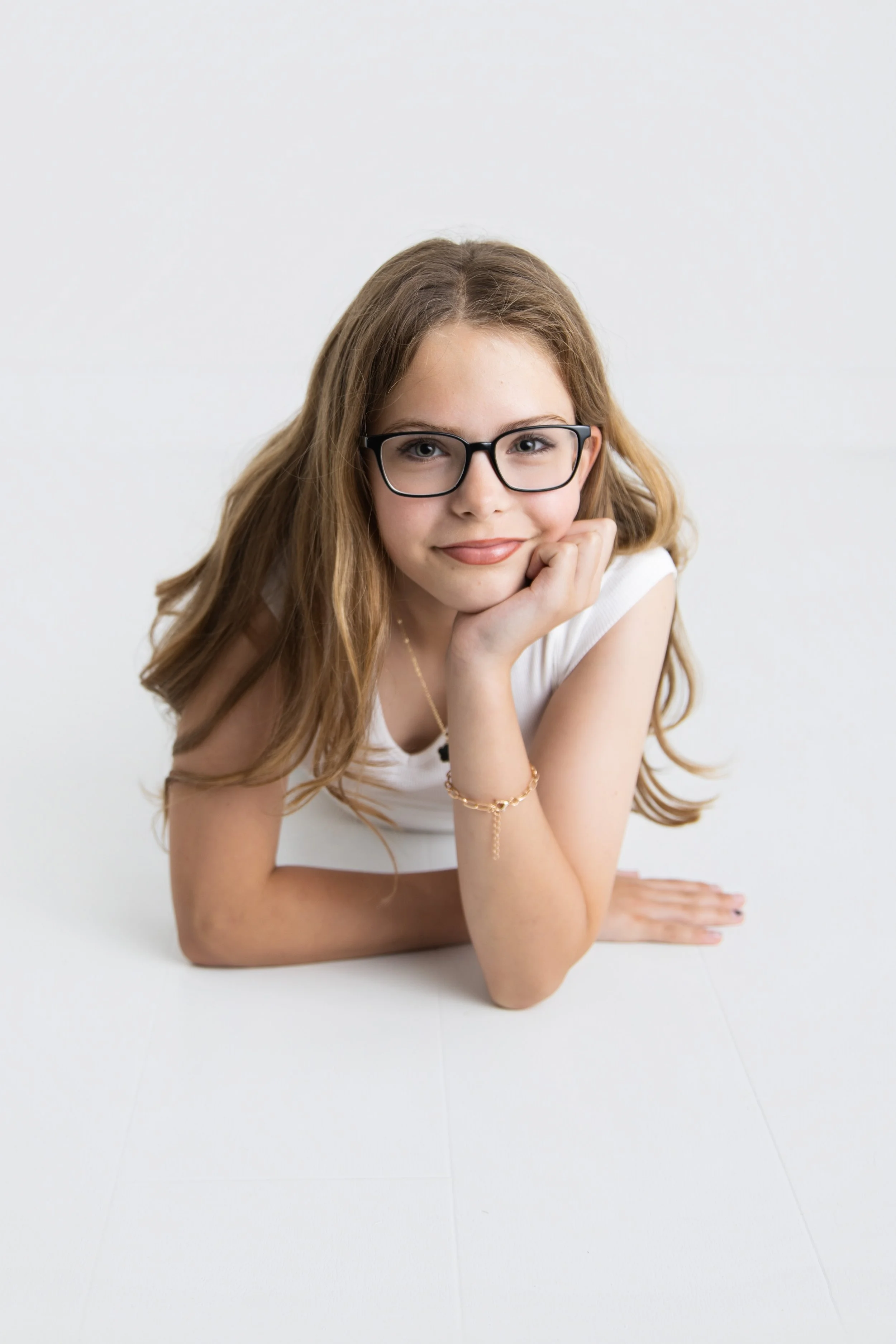Young girl with brown hair and glasses lying on her stomach on a white surface, supporting her chin with one hand, wearing a white sleeveless top, a gold bracelet, and a delicate necklace, smiling at the camera.
