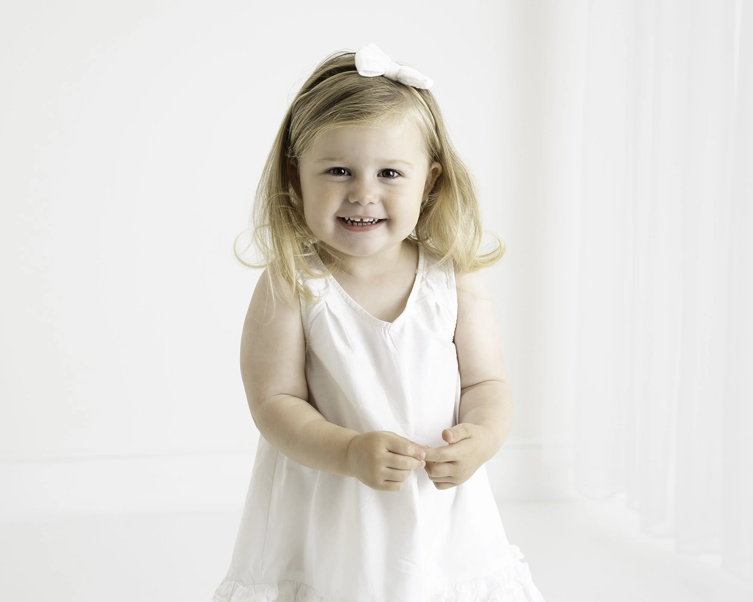 A young girl with blonde hair, wearing a white dress and a white bow headband, smiling at the camera in a bright, white room.