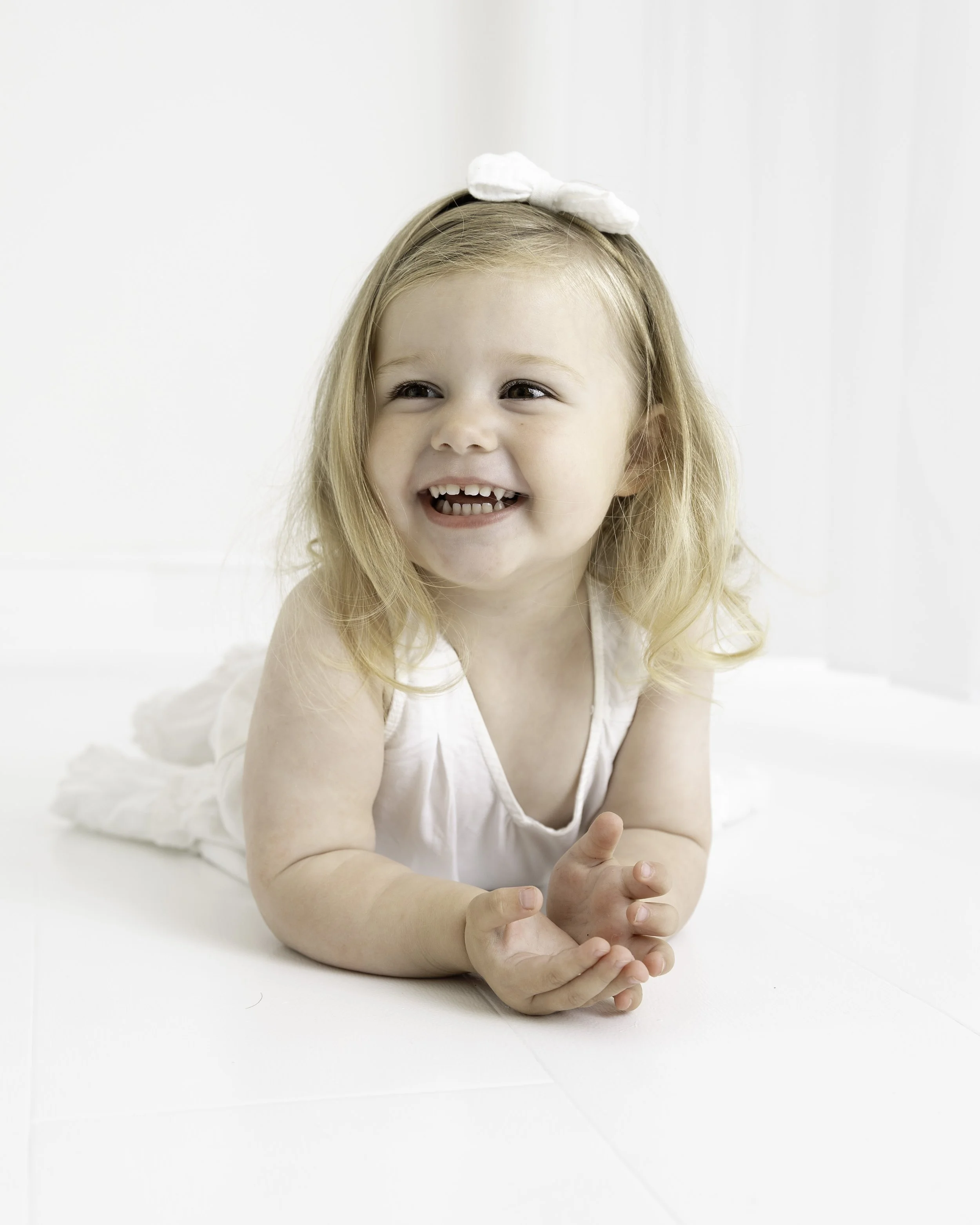 A smiling young girl with missing front teeth, lying on her stomach on a white surface, with a white bow in her hair, wearing a white sleeveless dress.