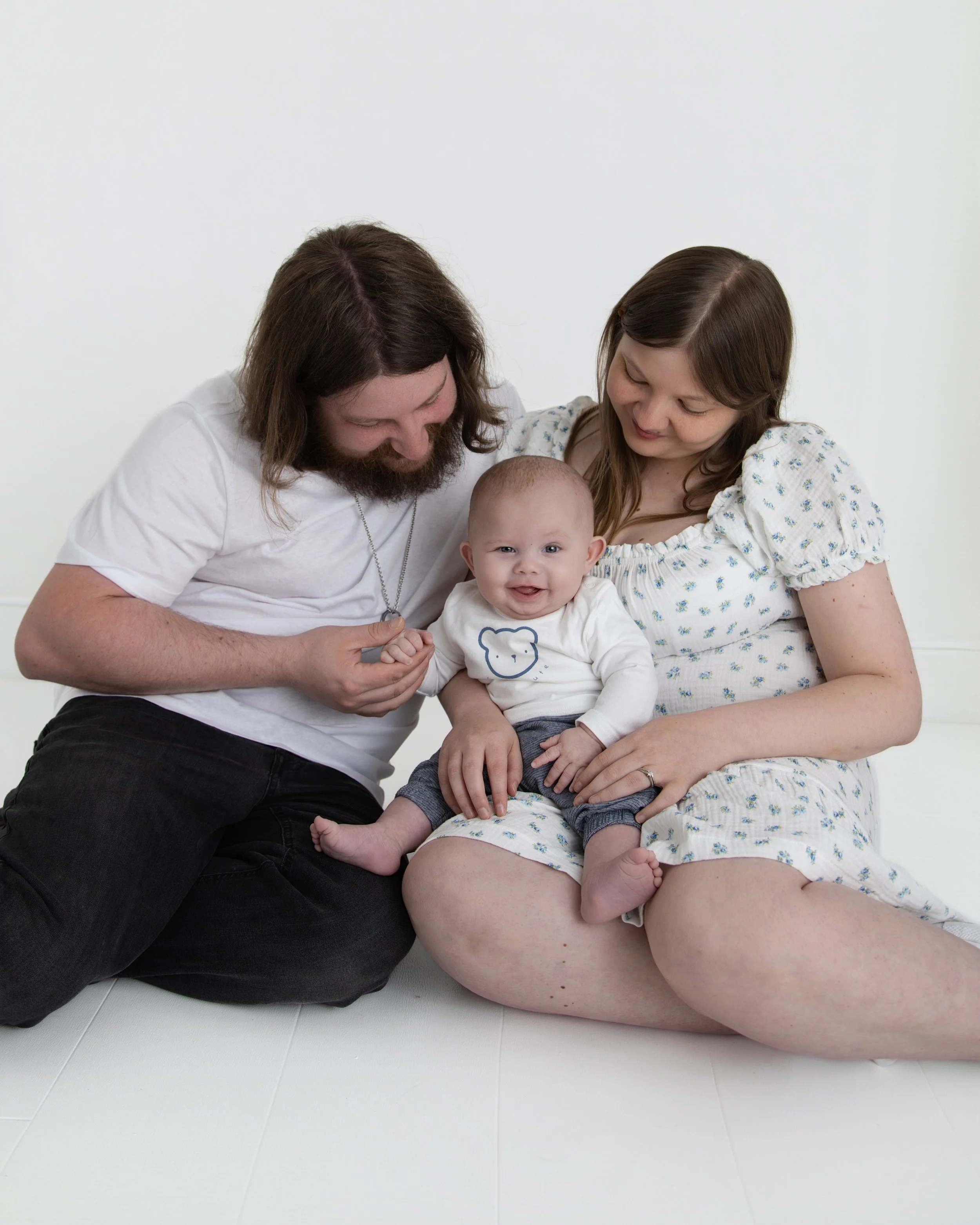 A family of three sitting on the floor, looking at a smiling baby who is sitting on the mother's lap. The father and mother are smiling at the baby.