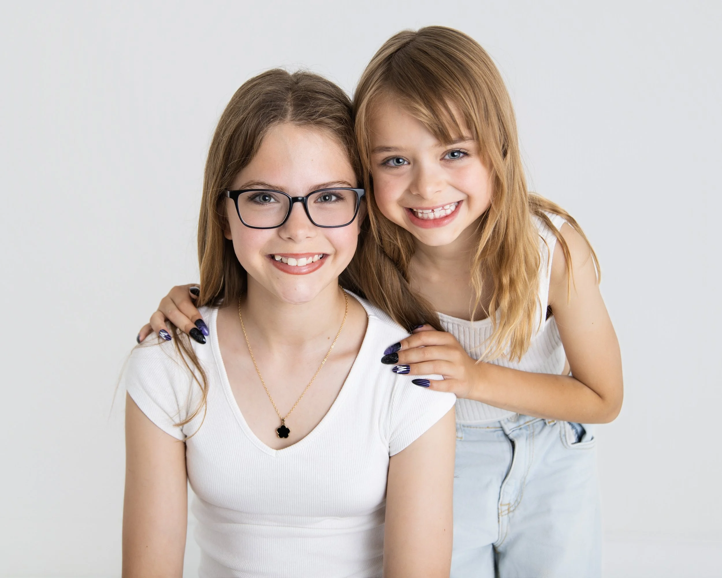 A woman with glasses and a young girl with blonde hair smiling together against a plain white background. The young girl is sitting behind the woman with her hand on the woman's shoulder.