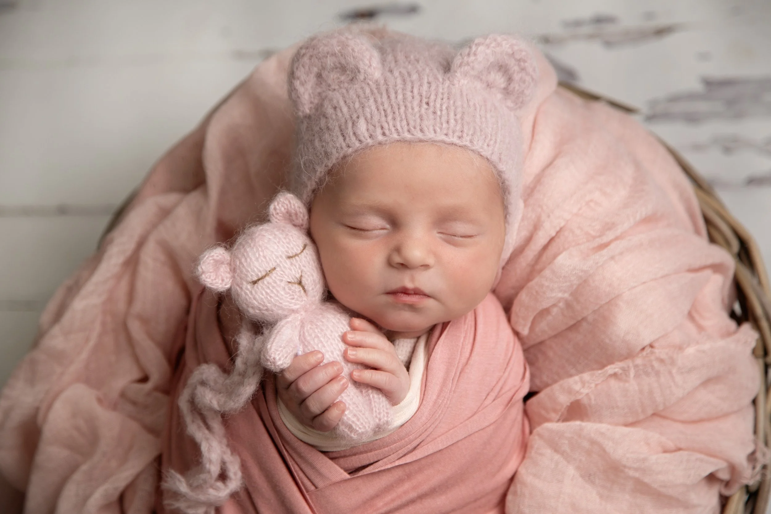 A sleeping baby wearing a pink knit hat with bear ears, holding a pink knitted bear plush toy, wrapped in pink blankets in a basket.