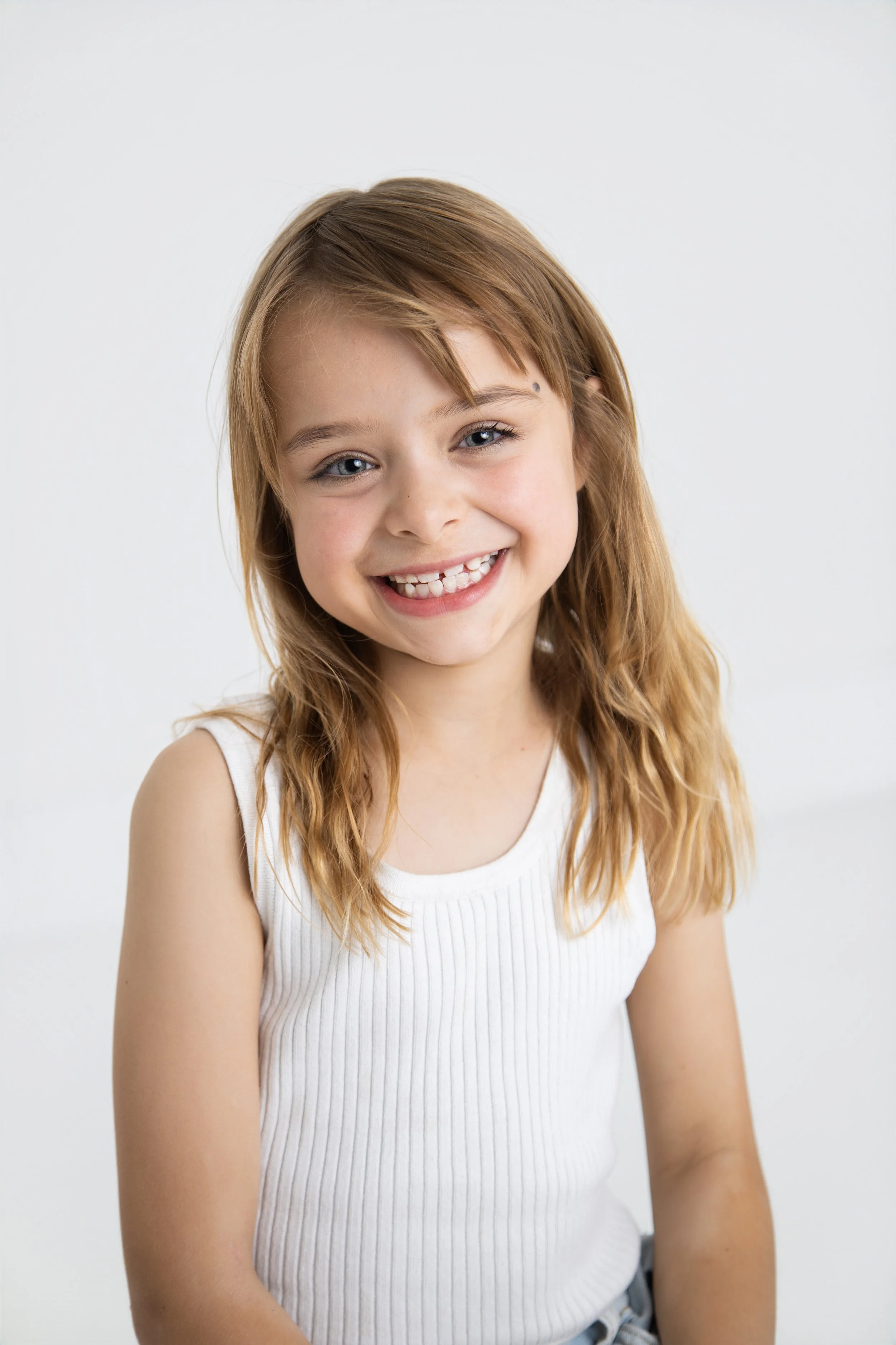 Smiling girl with red hair and blue eyes wearing a white sleeveless top against a plain white background.